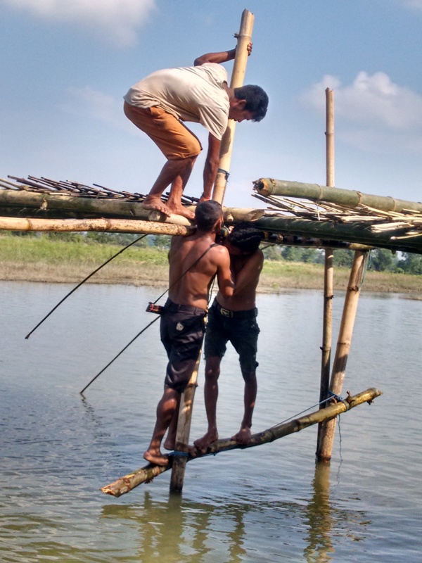 Two men stand on a bamboo raft over water, one man climbs a bamboo pole, the other supports him. Clear sky background.