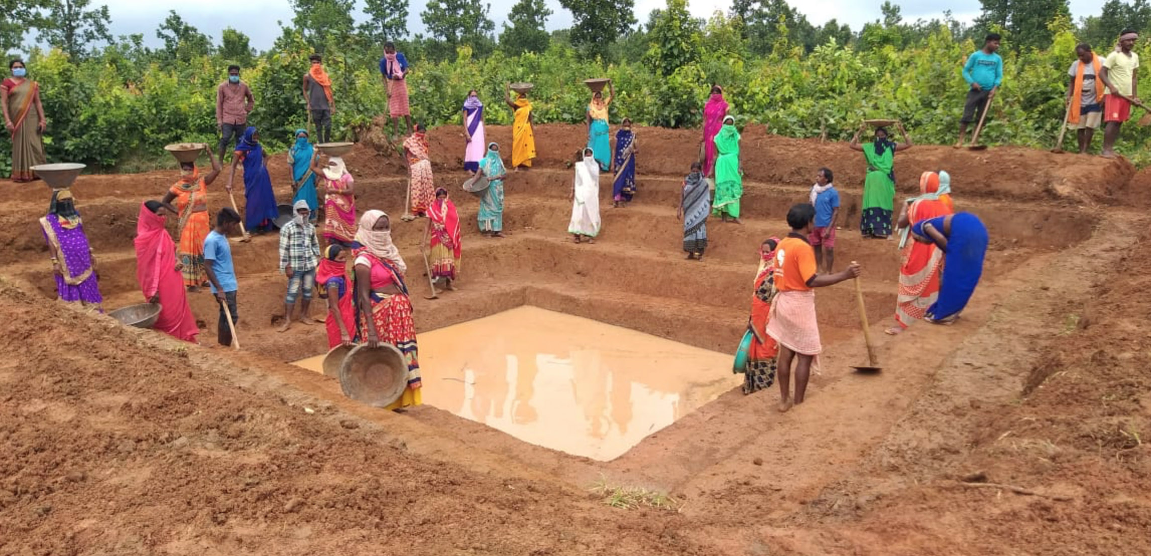 Group of people gathered around a large, rectangular waterhole in a rural area, some standing and some sitting.