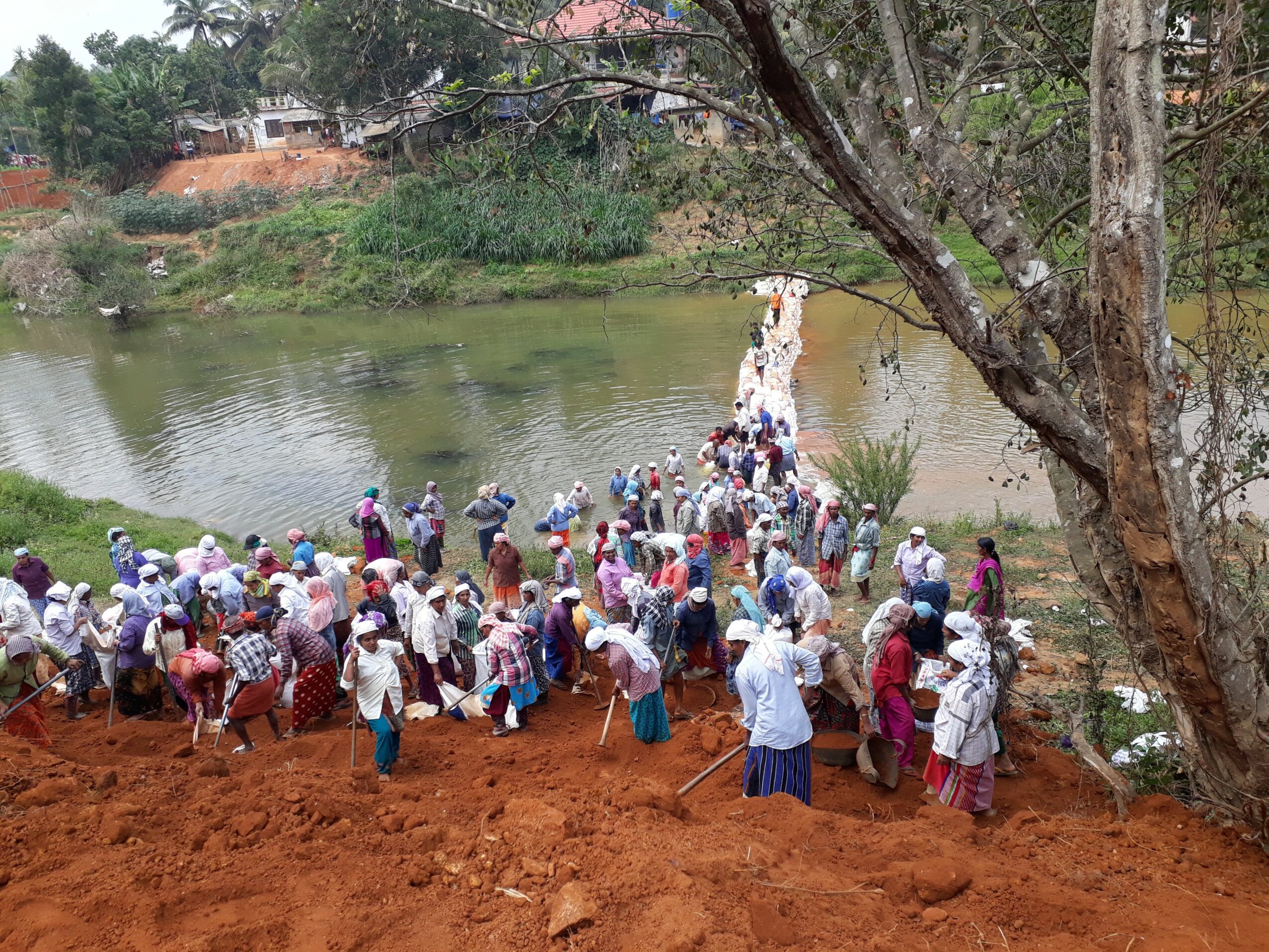 Large group of people gathered near a riverbank, some crossing a small bridge, with trees and buildings in the background.