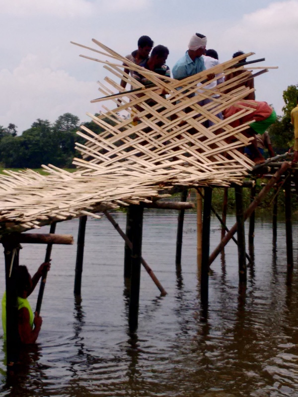 People standing on a bamboo bridge over water, constructing or repairing it, with trees and sky in the background.
