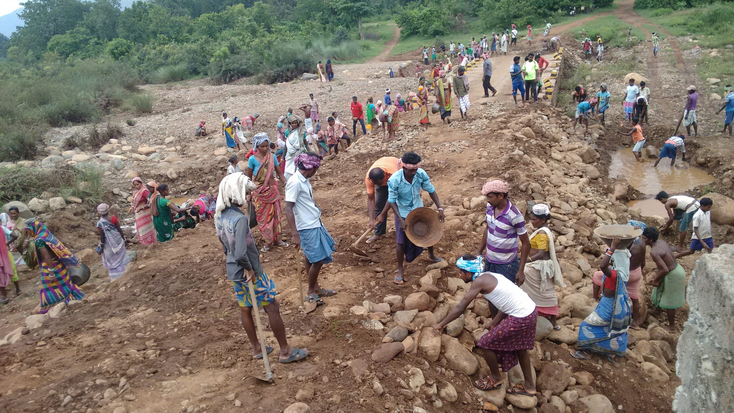 Group of people working on a large outdoor excavation site with dirt and rocks, surrounded by greenery and trees.