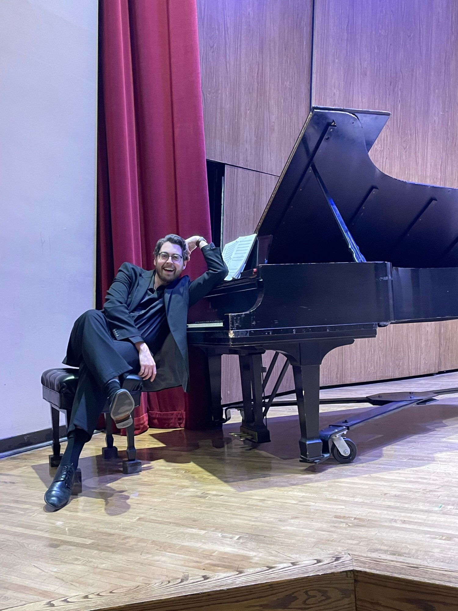 Person standing next to a grand piano on a wooden floor with red curtains in the background.