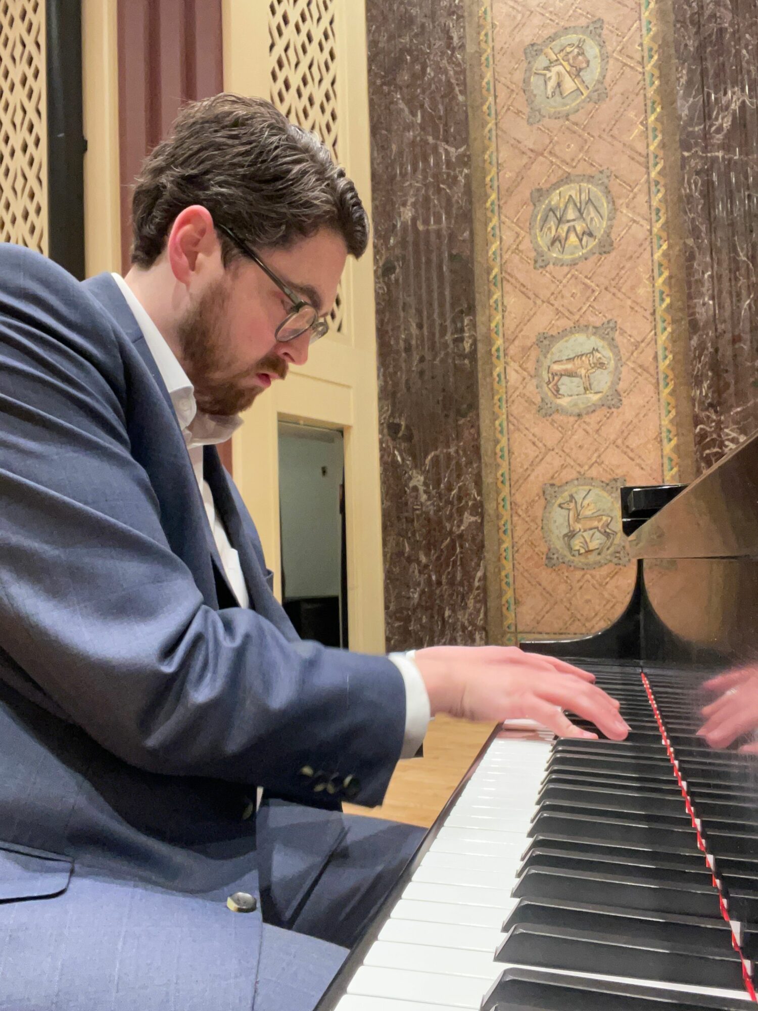 Person playing piano, wearing glasses and a suit, in a room with decorative wall and wooden furniture.