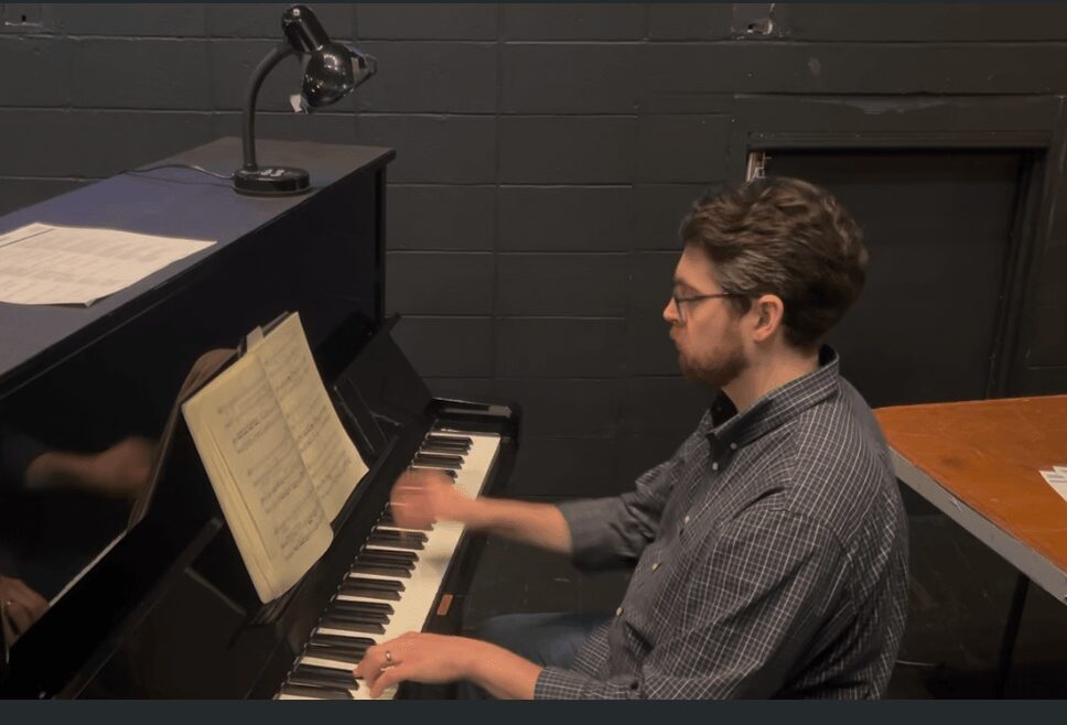 Young man playing piano with sheet music, in a room with dark walls and a desk nearby.