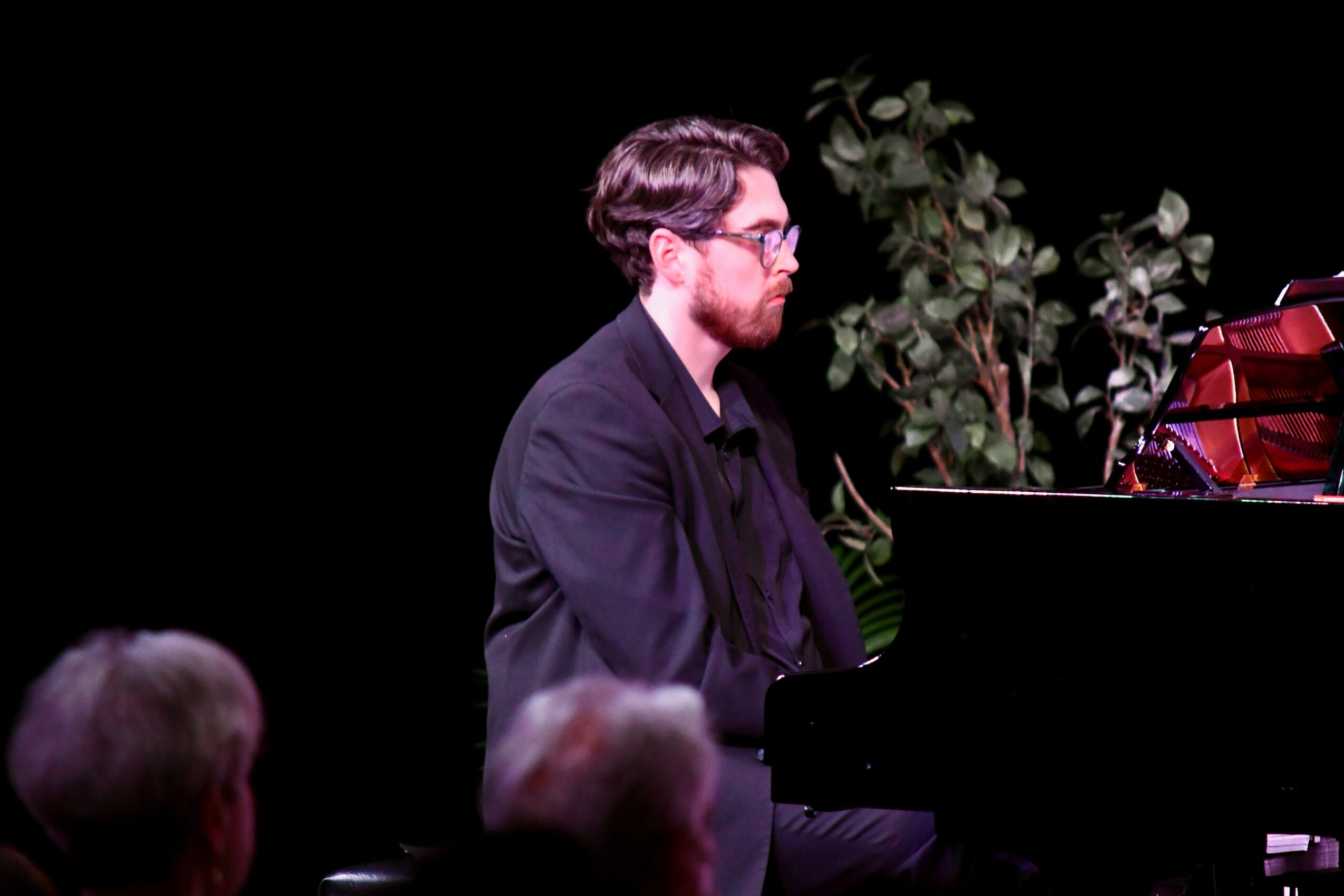 Man with glasses and beard playing piano on stage with audience watching, a plant in the background.