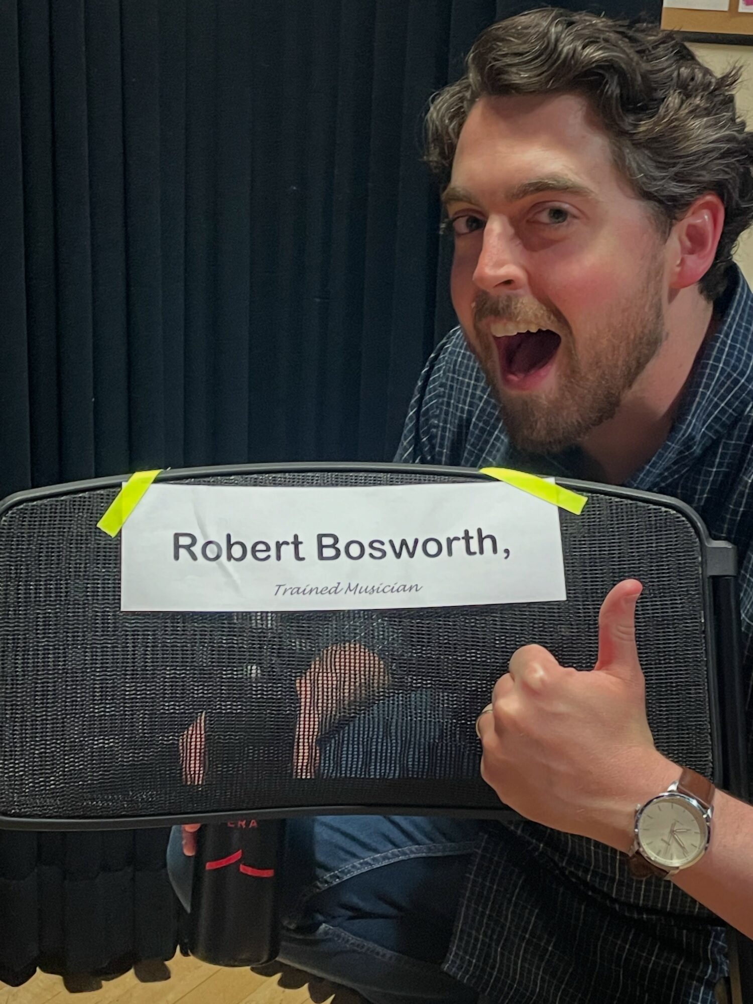 Man with curly hair and beard giving a thumbs-up, holding a sign with name and title, smiling, in front of a dark curtain.