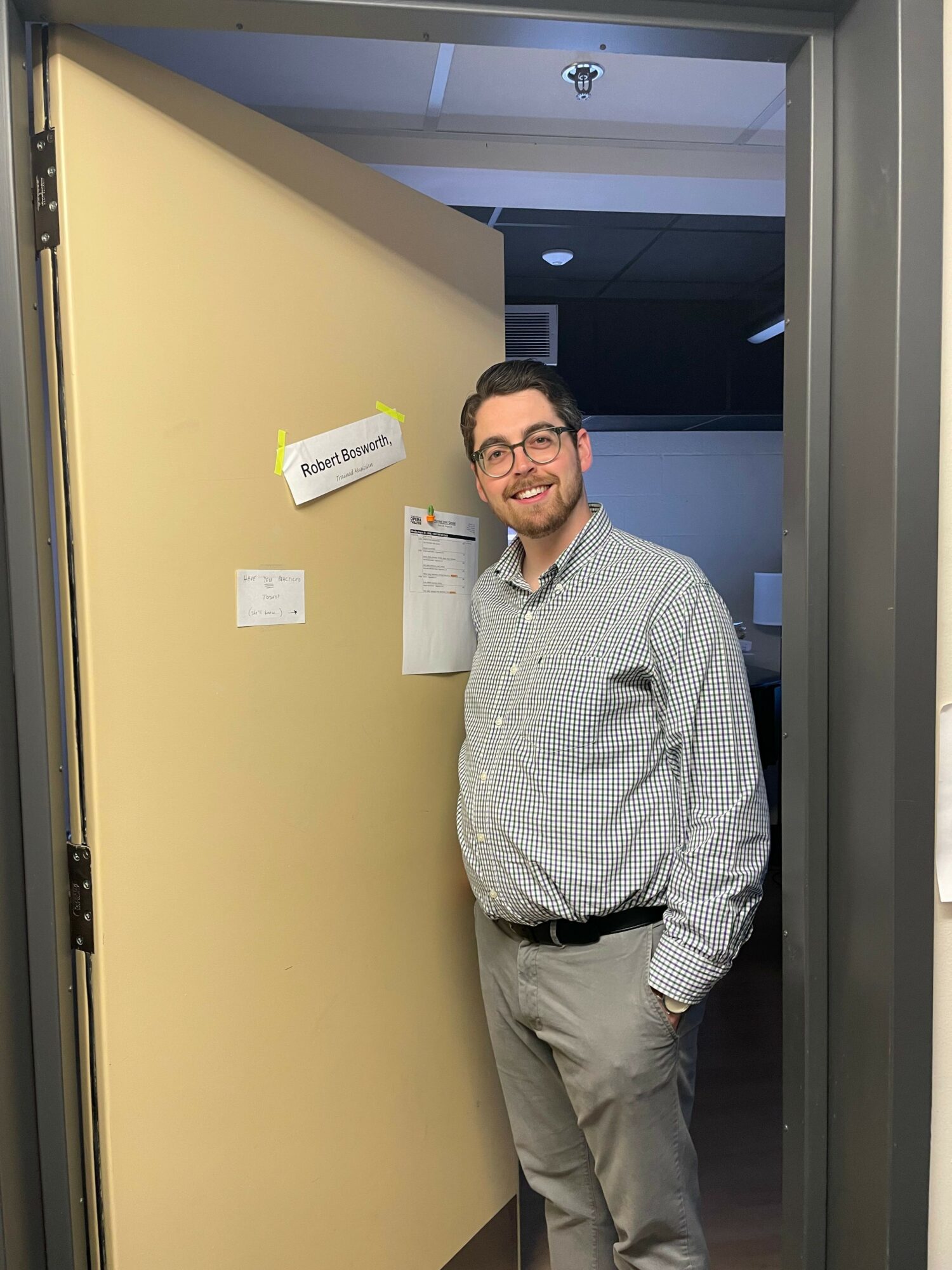 Man with glasses and a checkered shirt standing in a doorway, smiling, with a beige wall behind him.