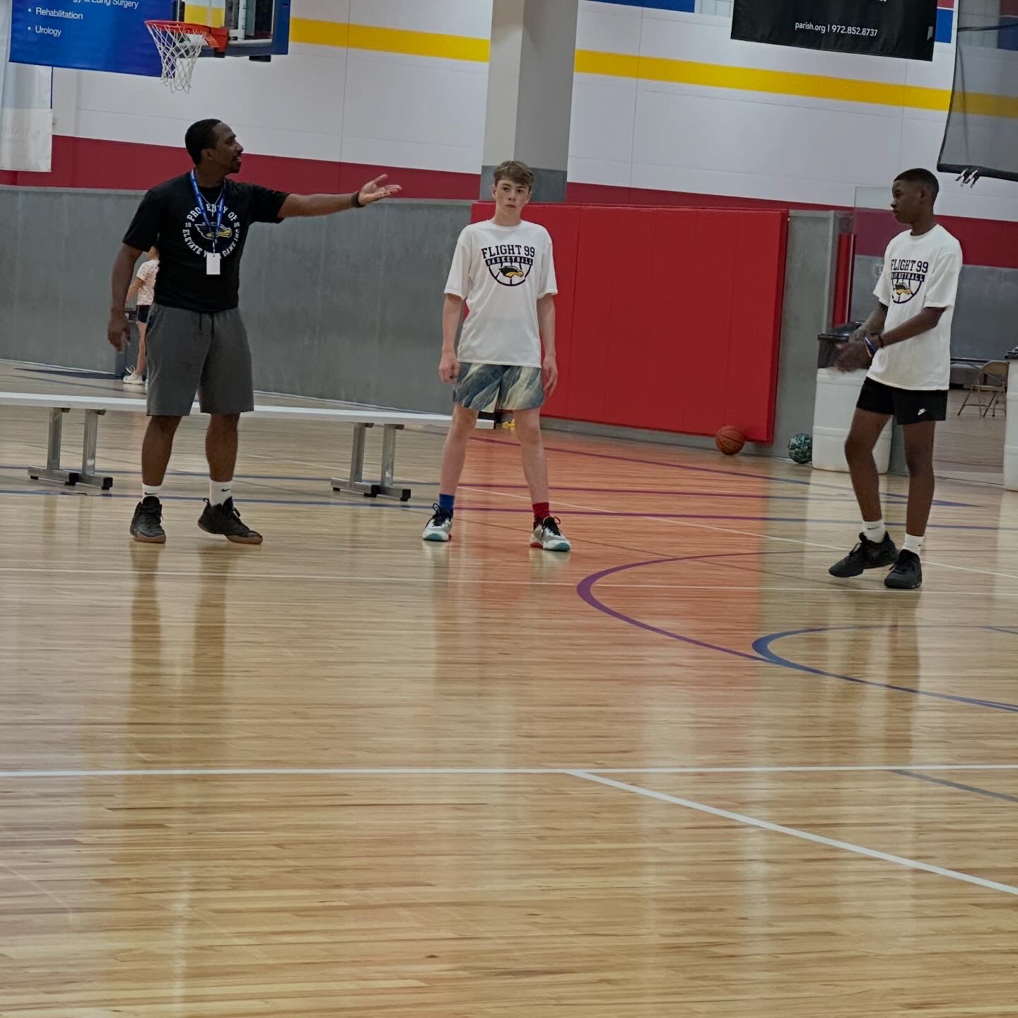 Three people in a gymnasium, one person appears to be coaching or instructing, with a basketball and gym equipment visible.