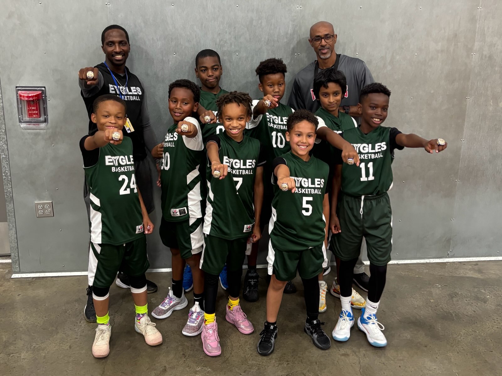 Group of young basketball players and coach posing indoors, some holding medals, smiling, against a plain wall.