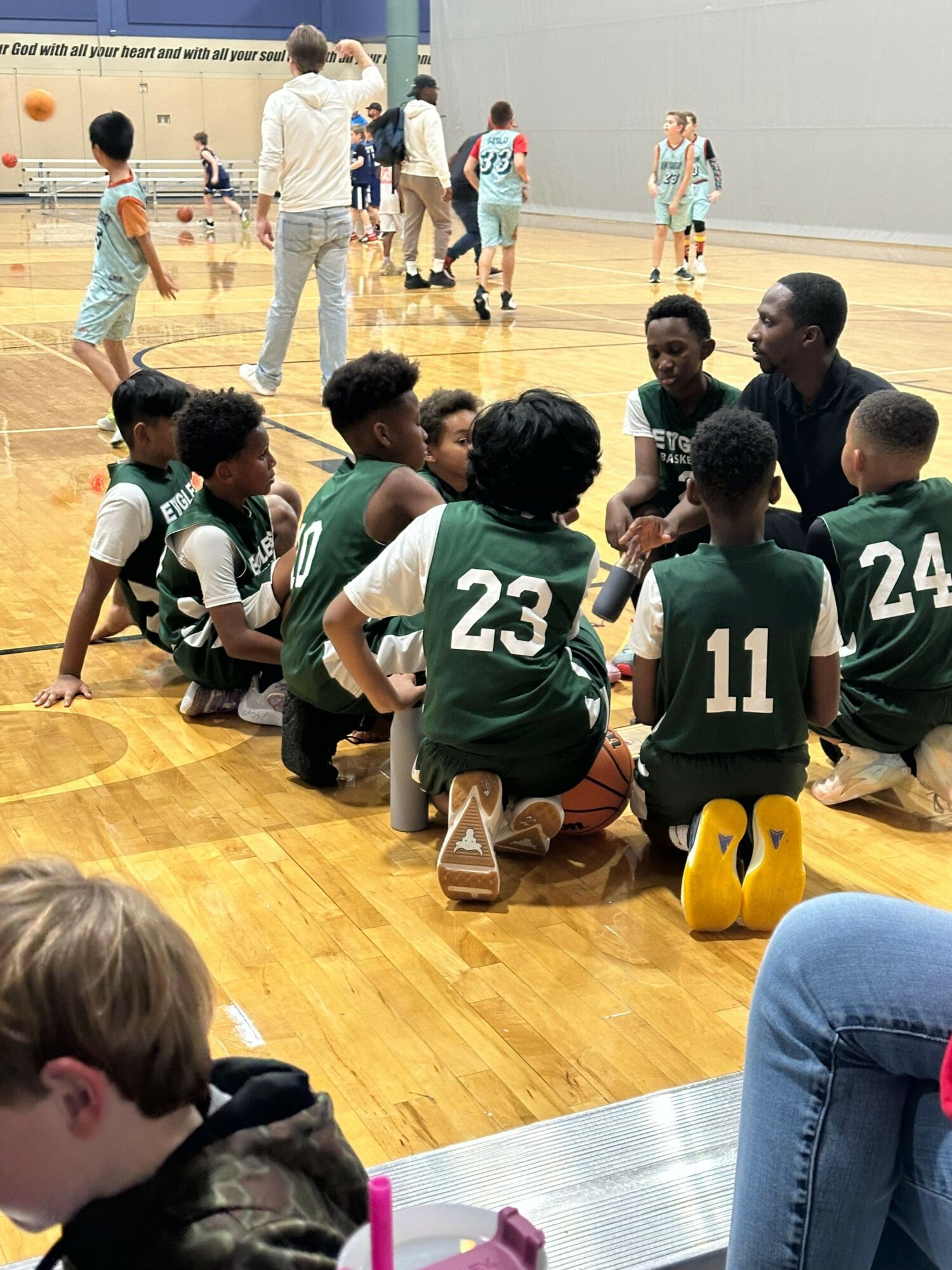 Group of young basketball players sitting on the floor during a game, with a referee and spectators in the background.