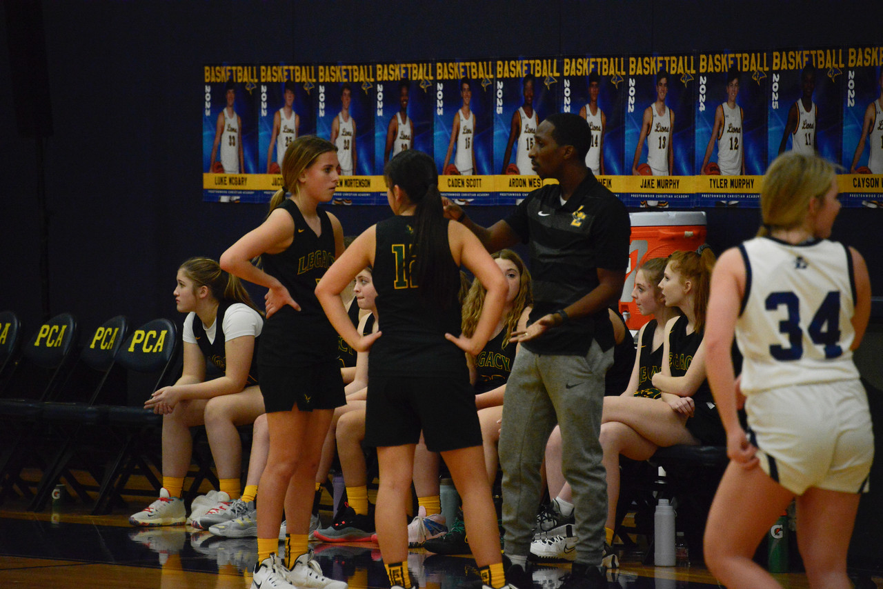 Basketball team huddle with coach during game, players seated and standing, gym background with banners, some players in white uniforms.