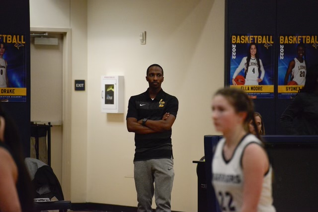 A man stands with arms crossed in a hallway, with posters on the wall and a girl in a sports uniform in foreground.