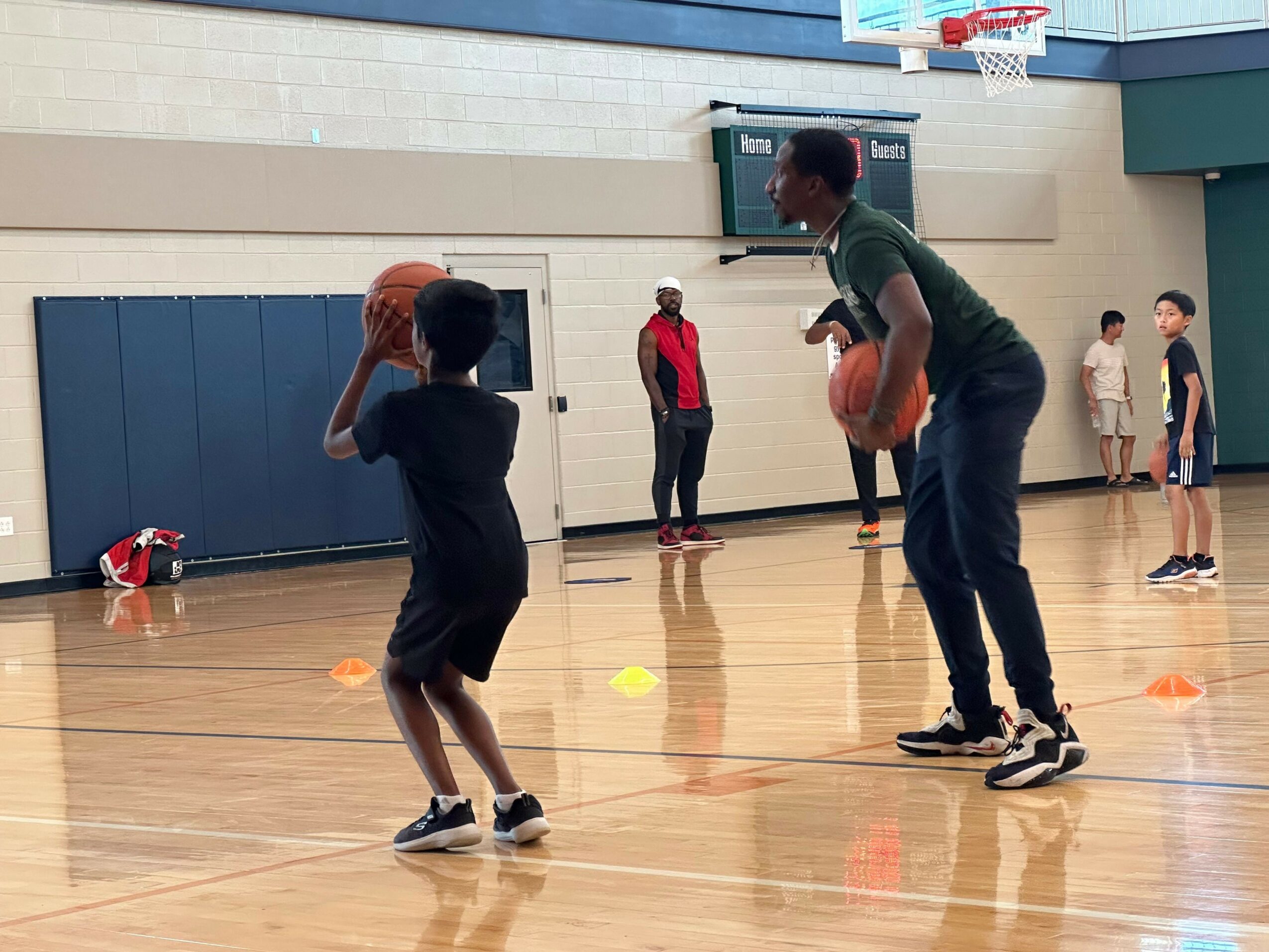 Two children playing basketball in a gym, one holding a ball, with other children and cones in the background.