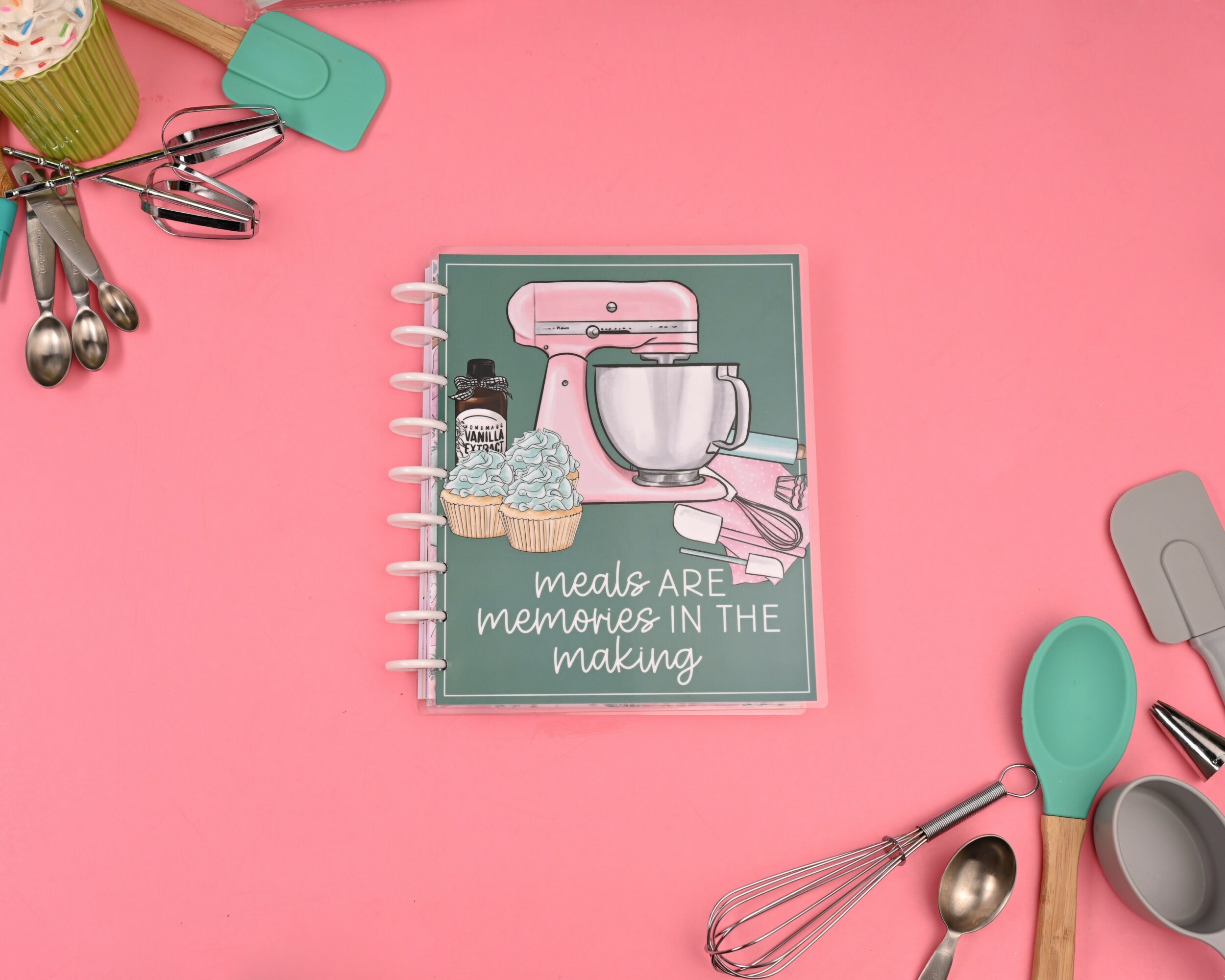 Pink background with a notebook featuring a mixer, bowl, and utensils, surrounded by kitchen tools and utensils.