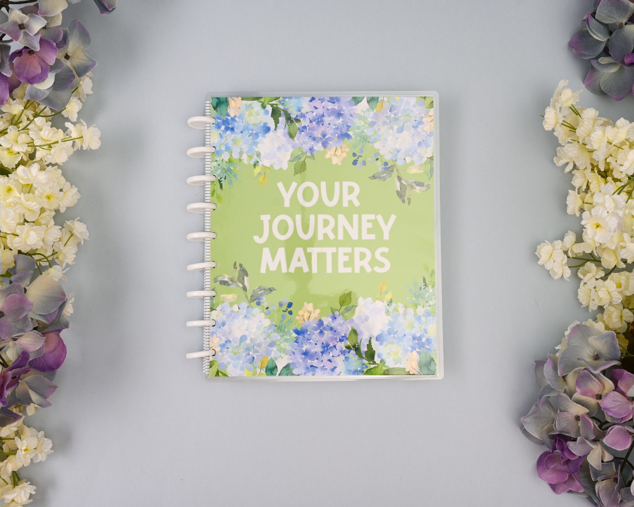 A spiral-bound notebook with a floral cover and the text 'Your Journey Matters' in the center, surrounded by flowers.