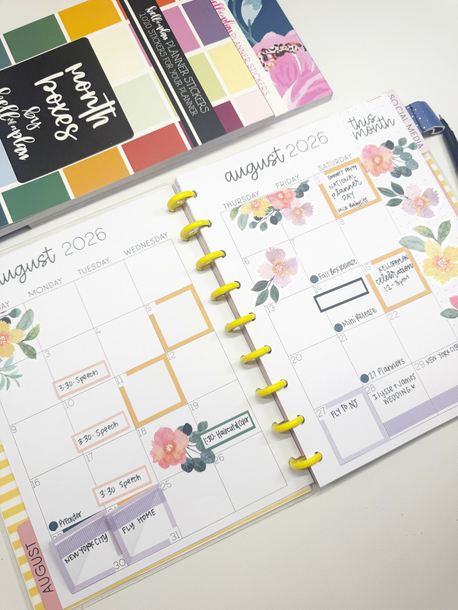Open planner showing August 2026 calendar pages with floral decorations and handwritten notes, with colorful sticker sheets above.