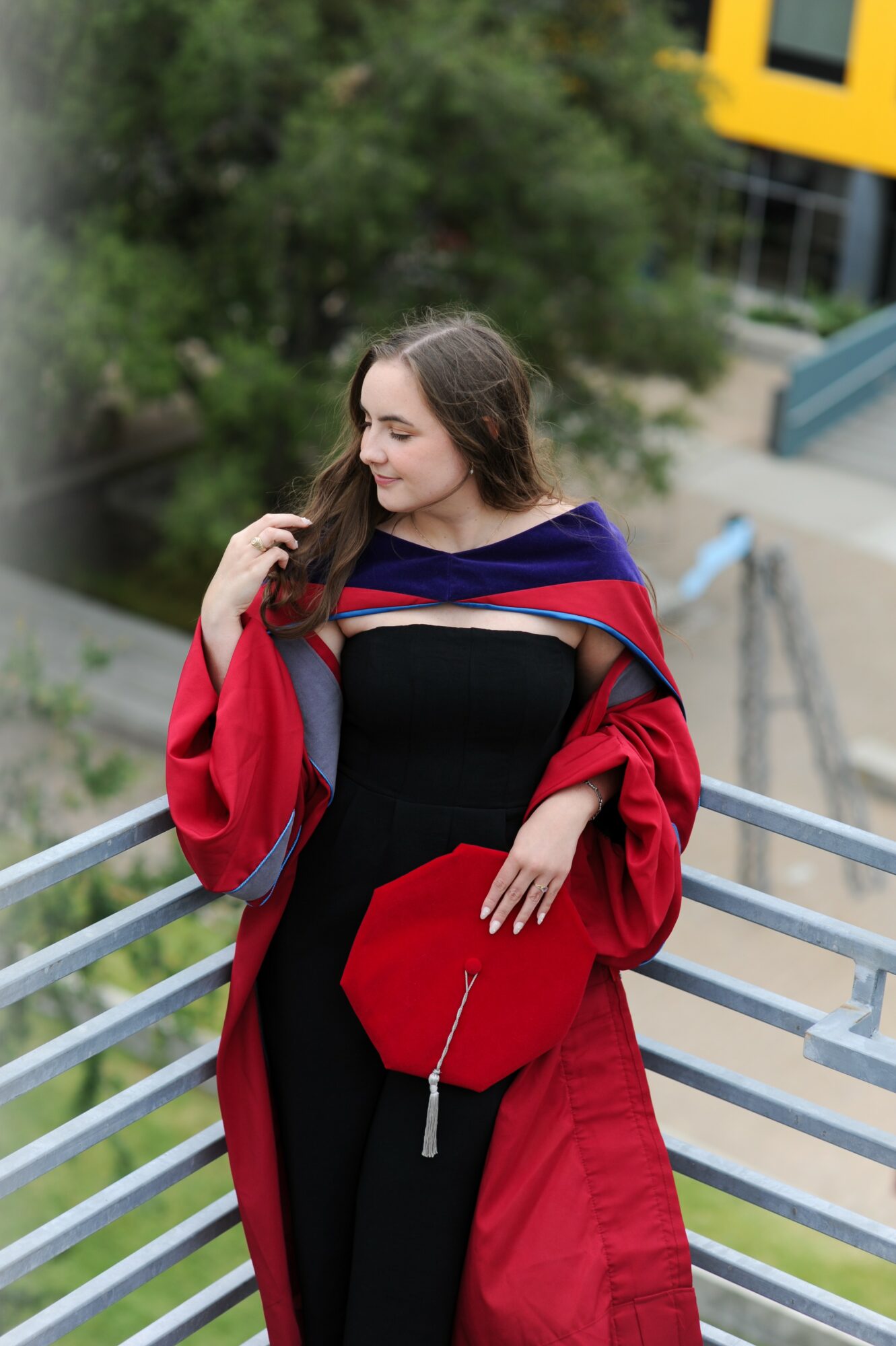 Young woman in a graduation gown holding a cap, standing outdoors on a metal railing, smiling, with trees and buildings in background.