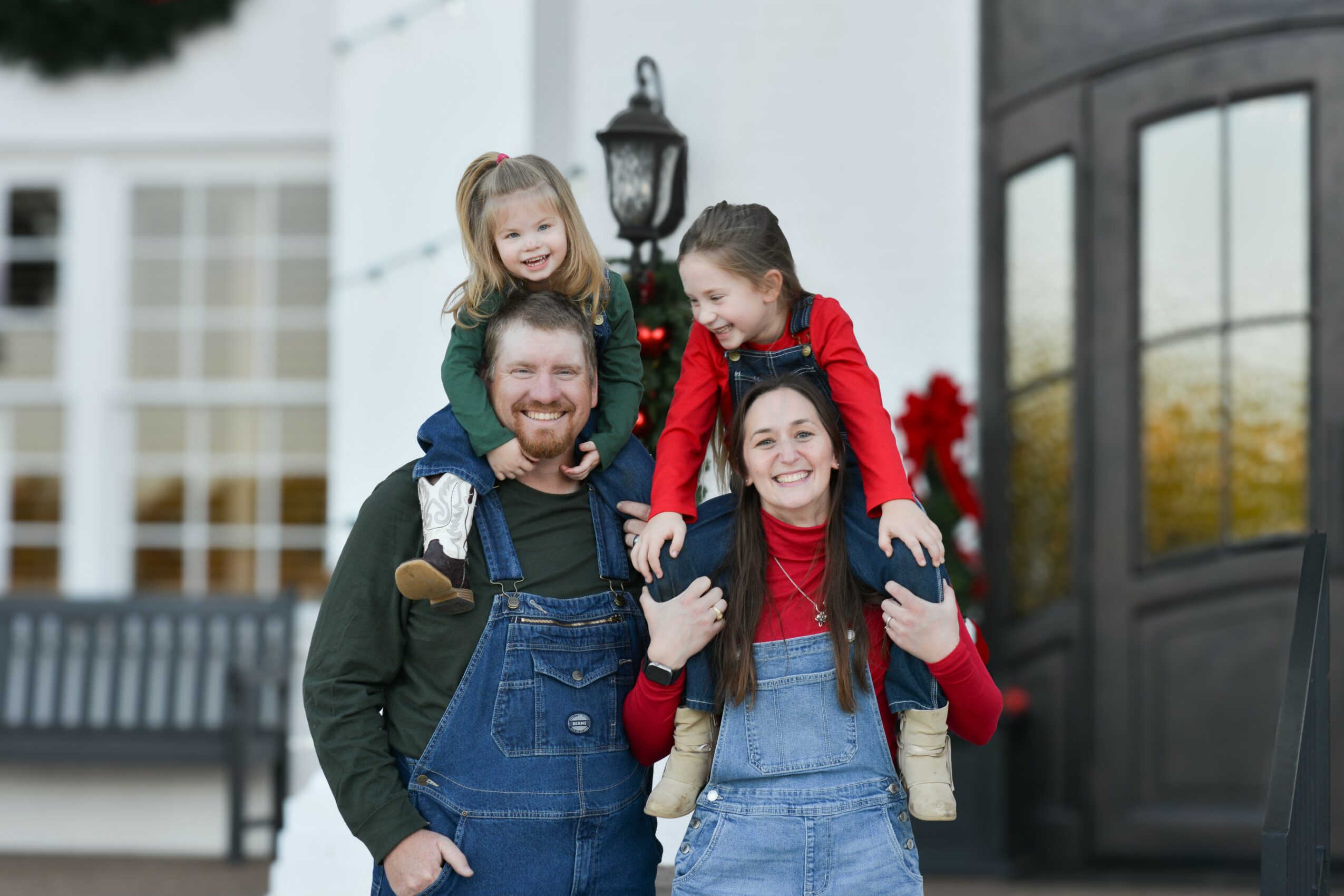 Family of four smiling outdoors, two children on parents' shoulders, in front of a white building with a bench and door.