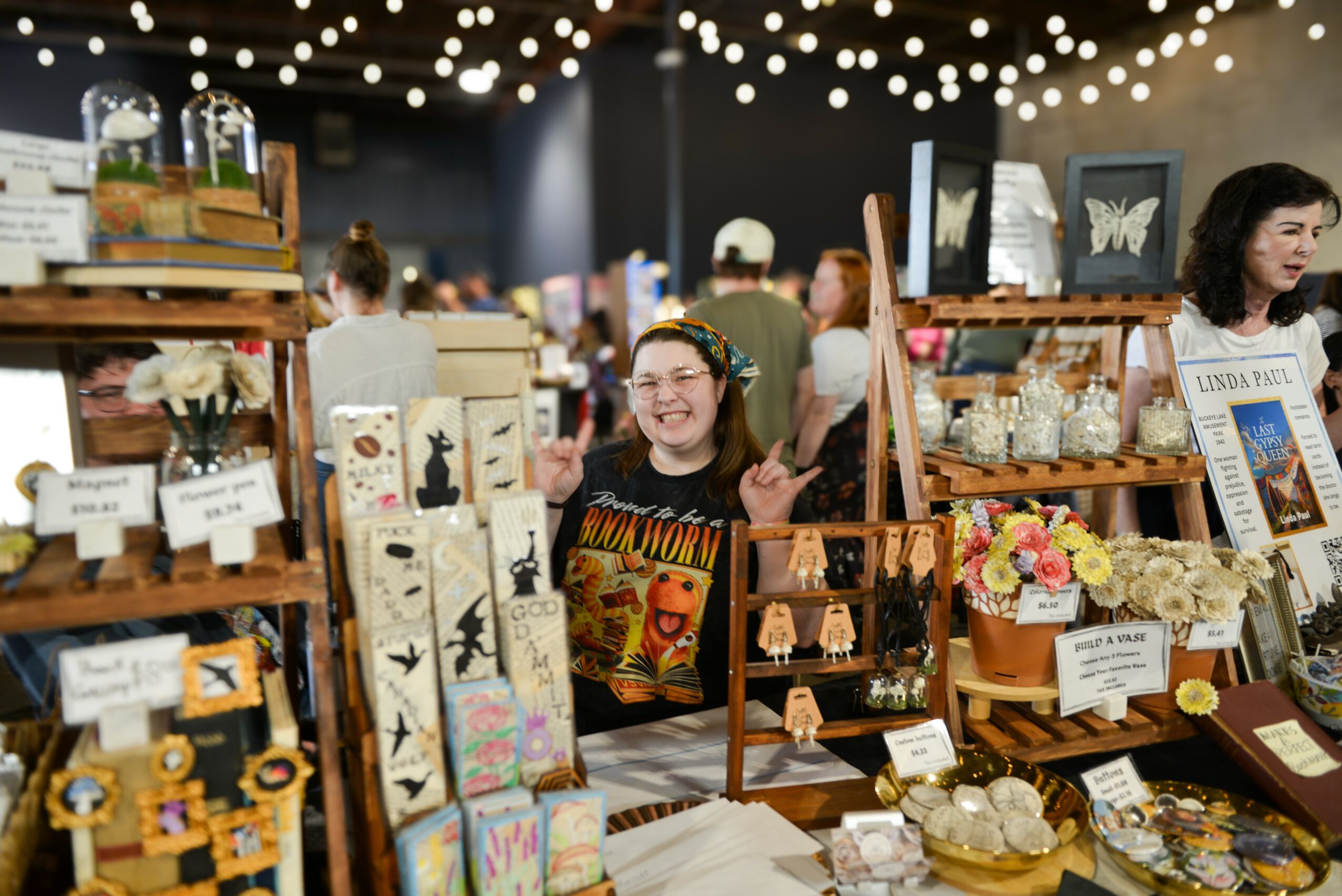 Girl smiling at a craft fair booth with jewelry and decorative items, other people browsing in background, string lights overhead.