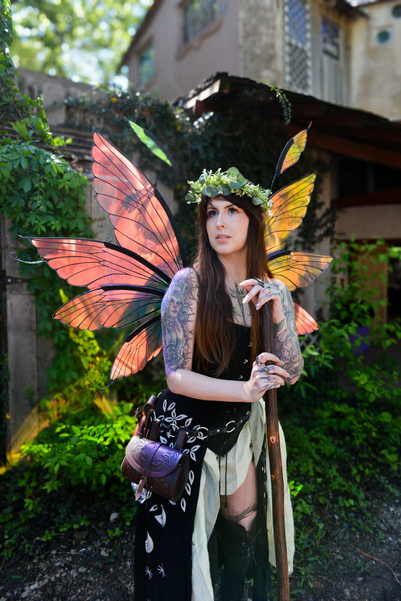 Woman dressed as a fairy with large pink and yellow wings, wearing a flower crown, standing outdoors with greenery.