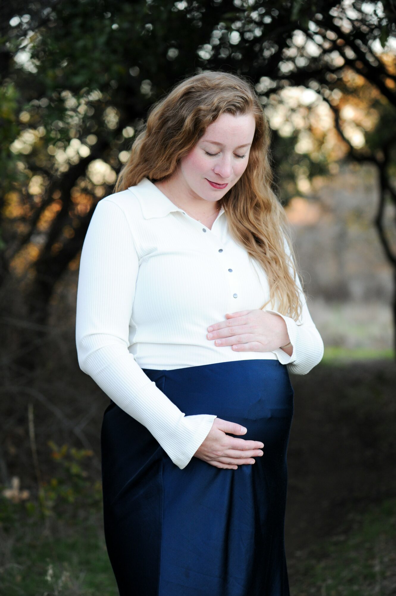 Pregnant woman with long hair in a white shirt and dark pants outdoors, holding her belly, trees in background.