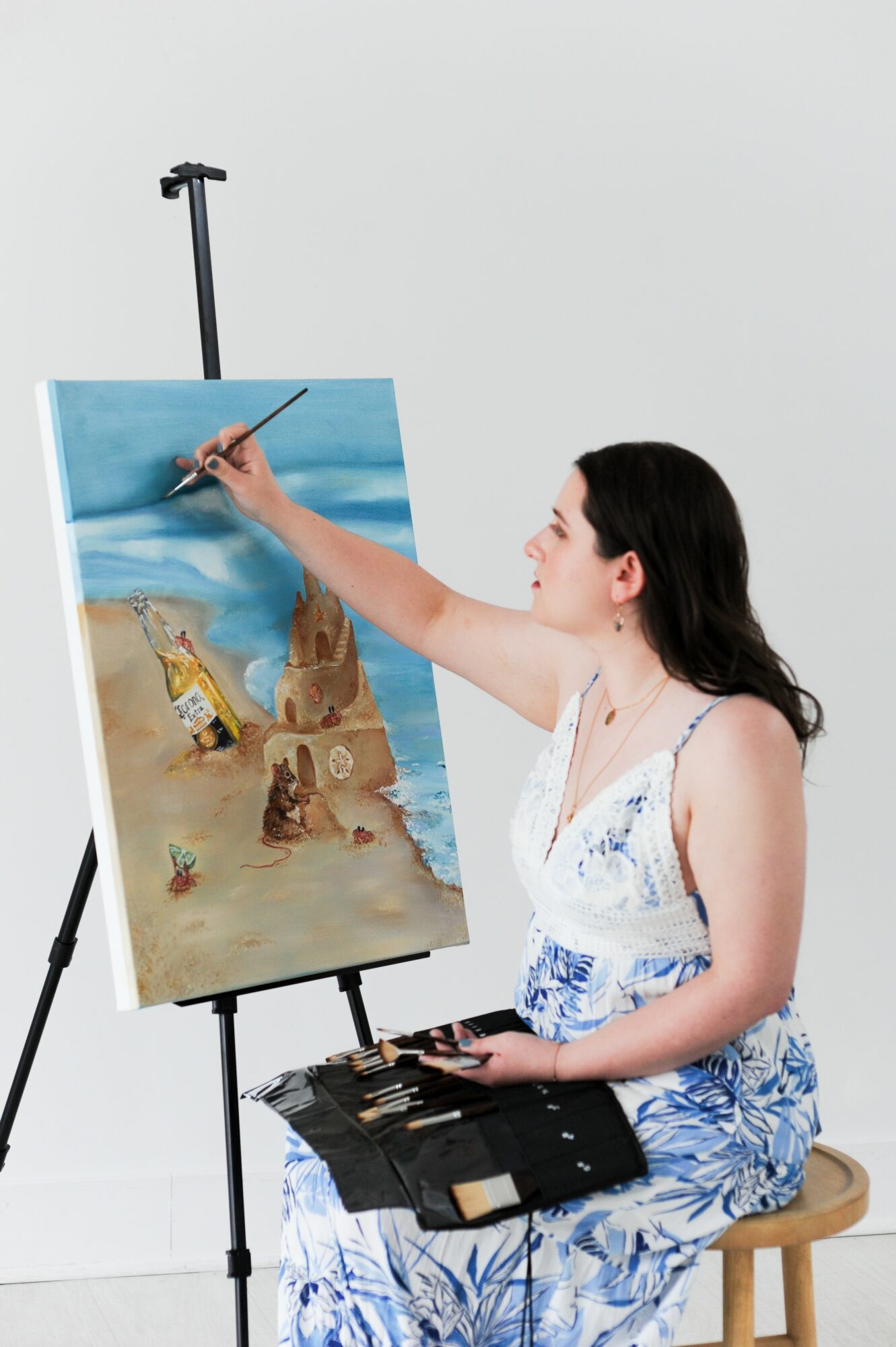 Woman painting on canvas with a seaside landscape, sitting on a stool, holding a palette, against a plain white background.