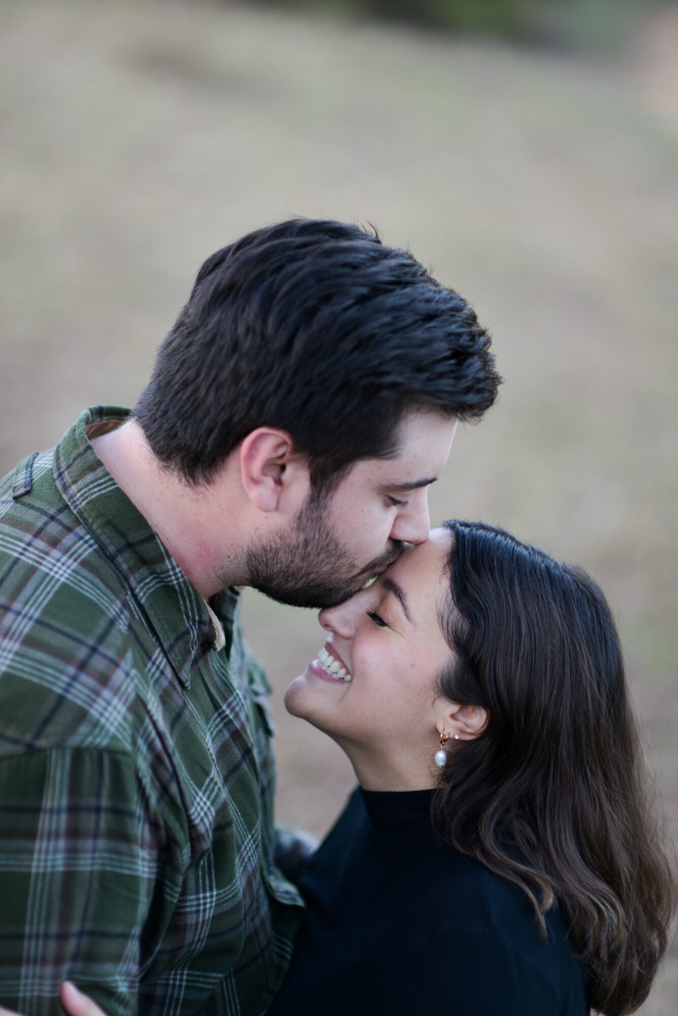 A man and woman face each other outdoors, smiling and touching foreheads, with a blurred natural background.