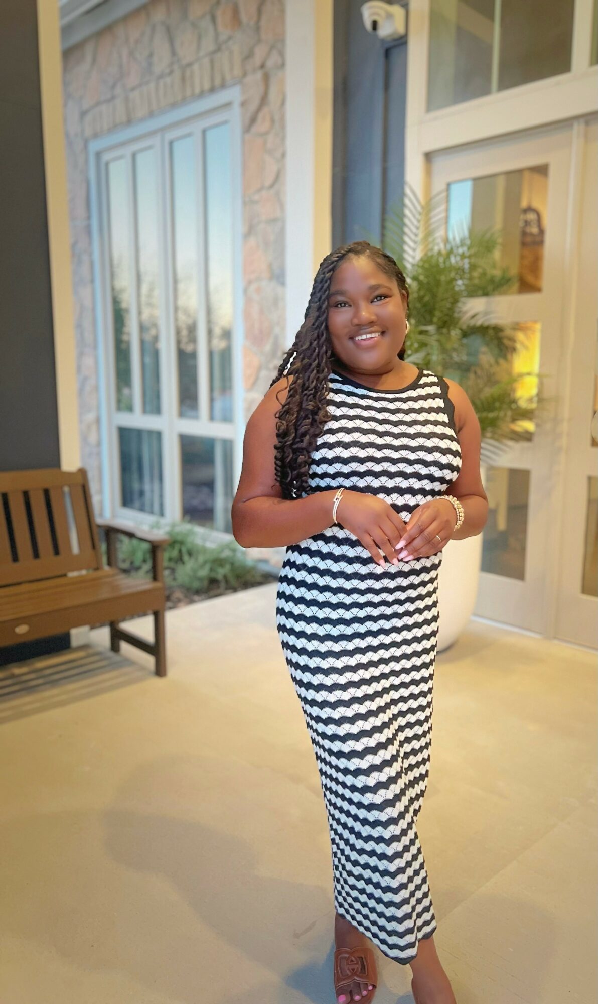 Smiling woman in a black and white striped dress standing outside near a building with large windows and a bench.