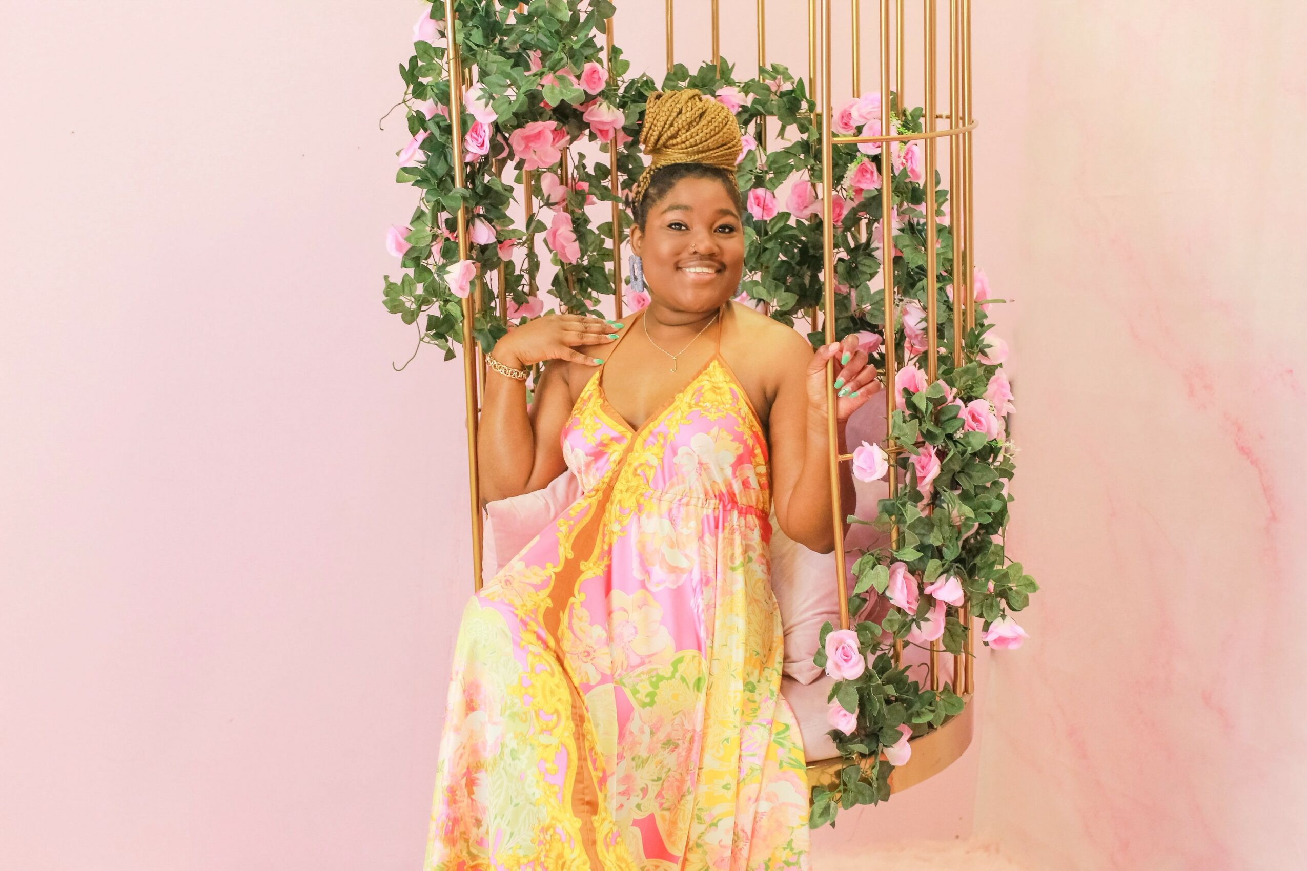Smiling woman in a colorful dress sitting on a swing decorated with pink and green flowers, against a pink background.