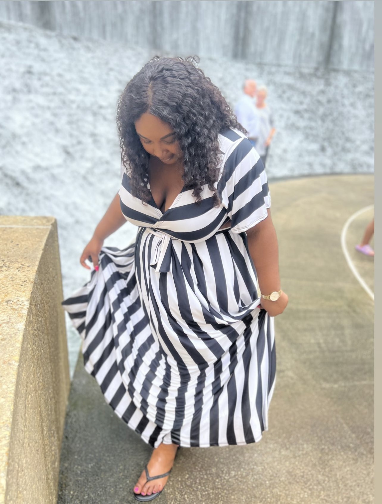 Woman in a black and white striped dress standing outdoors, looking down, with curly hair and sandals.