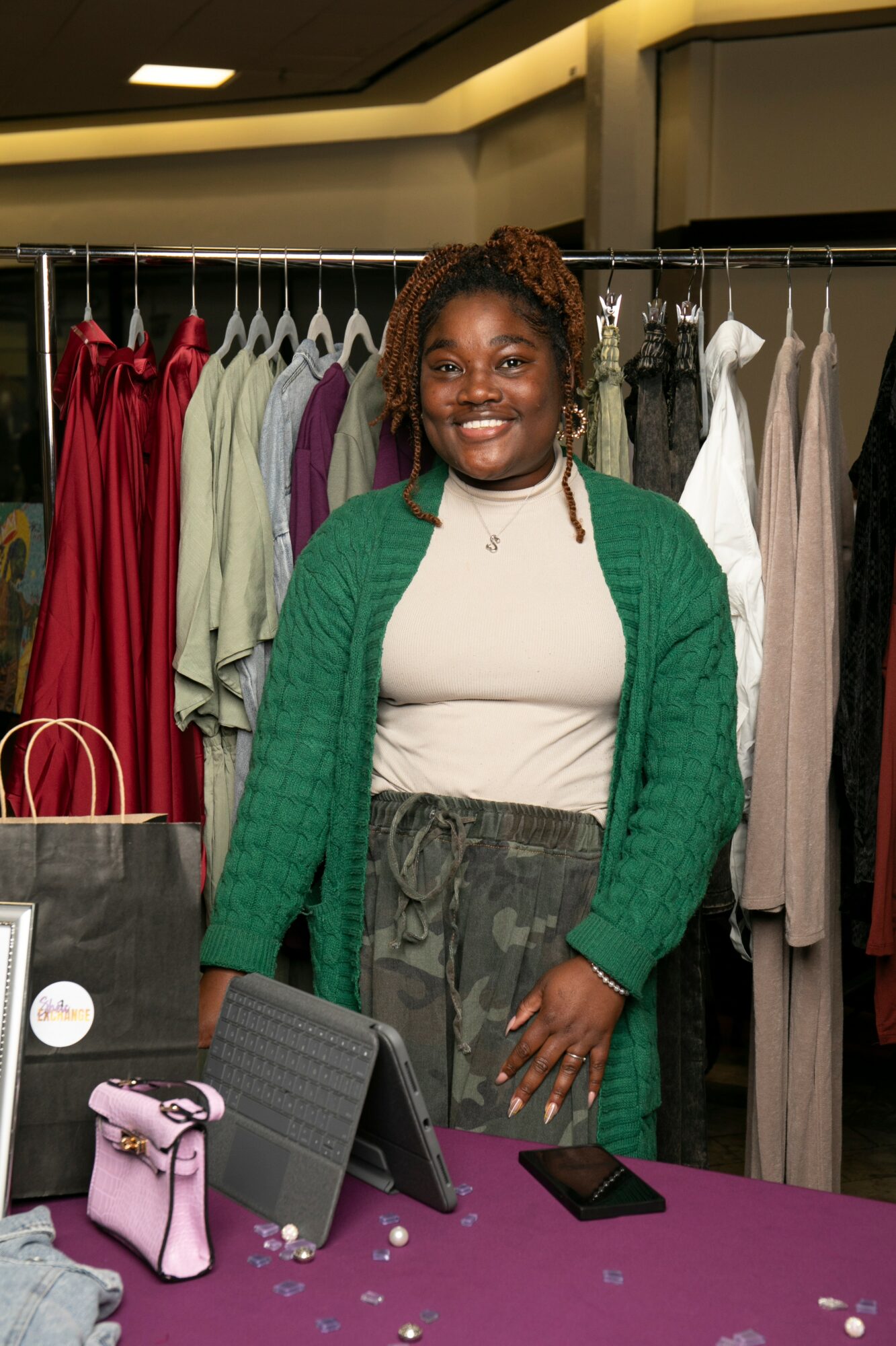 Smiling woman standing in front of clothing racks, wearing a green cardigan and white top, with a purple purse on table.