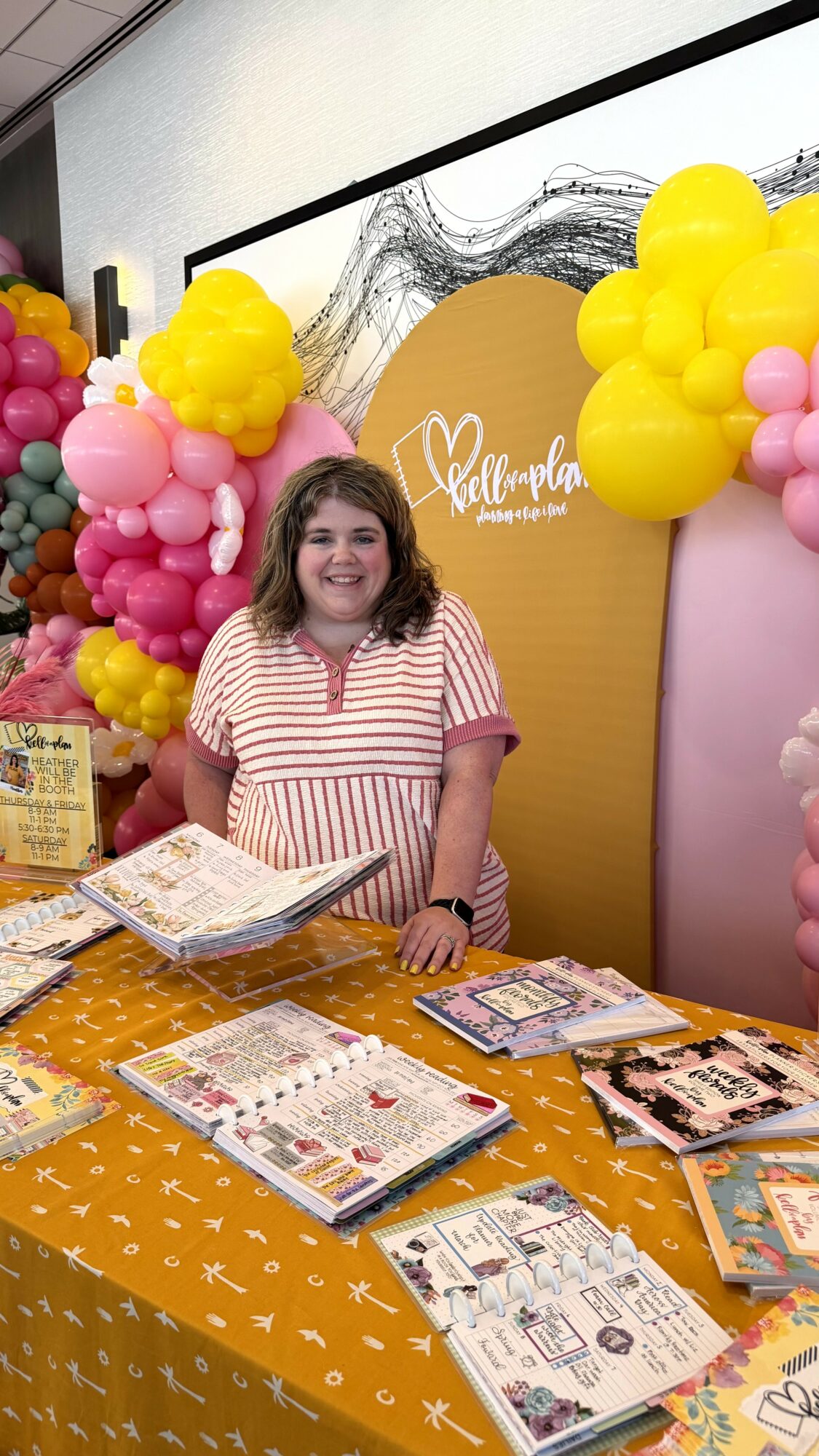 Smiling woman standing behind a table with magazines, decorated with pink, yellow, and purple balloons, and a yellow backdrop.
