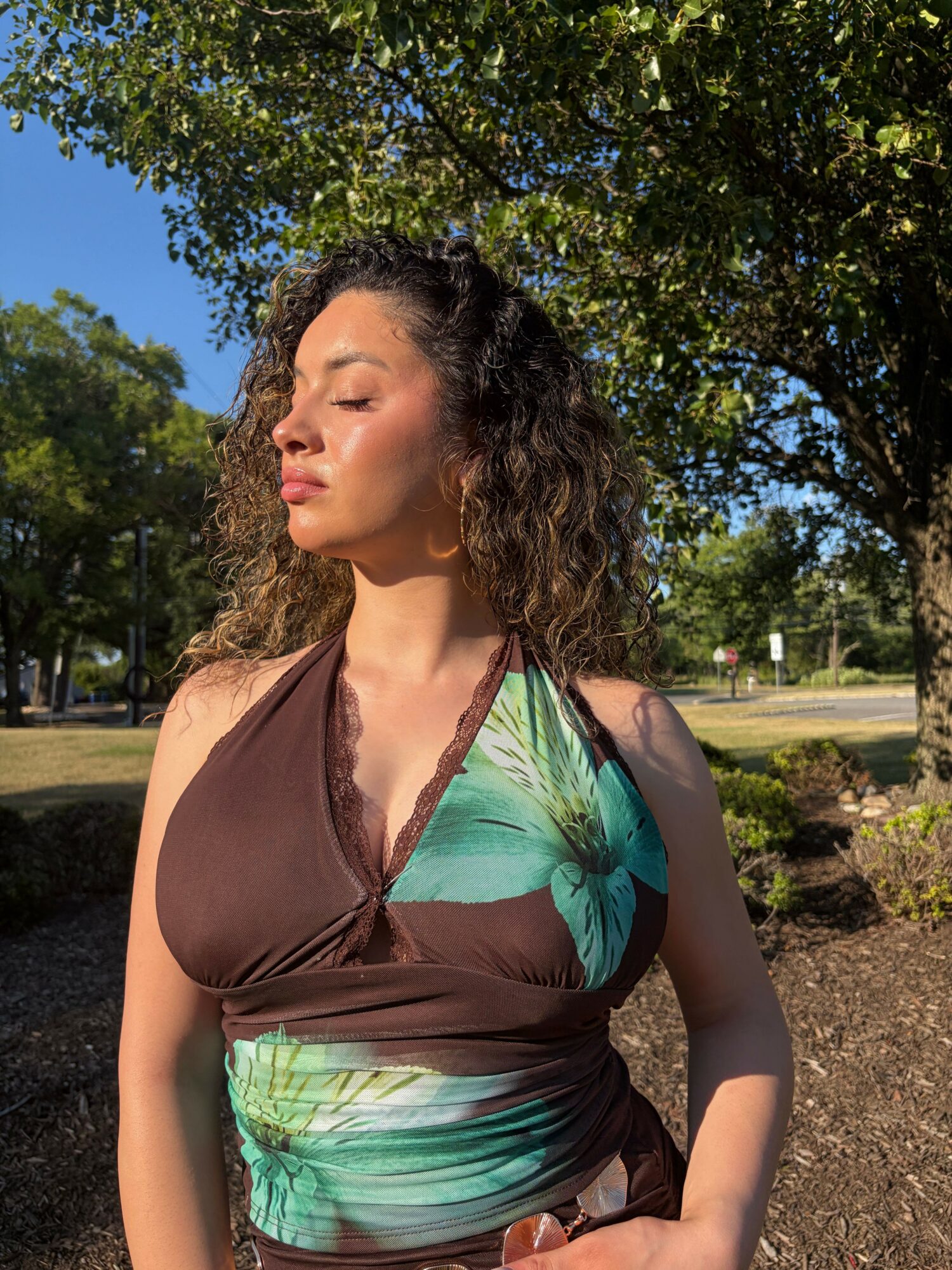 Woman with curly hair in a green and brown dress outdoors, trees and blue sky in background.