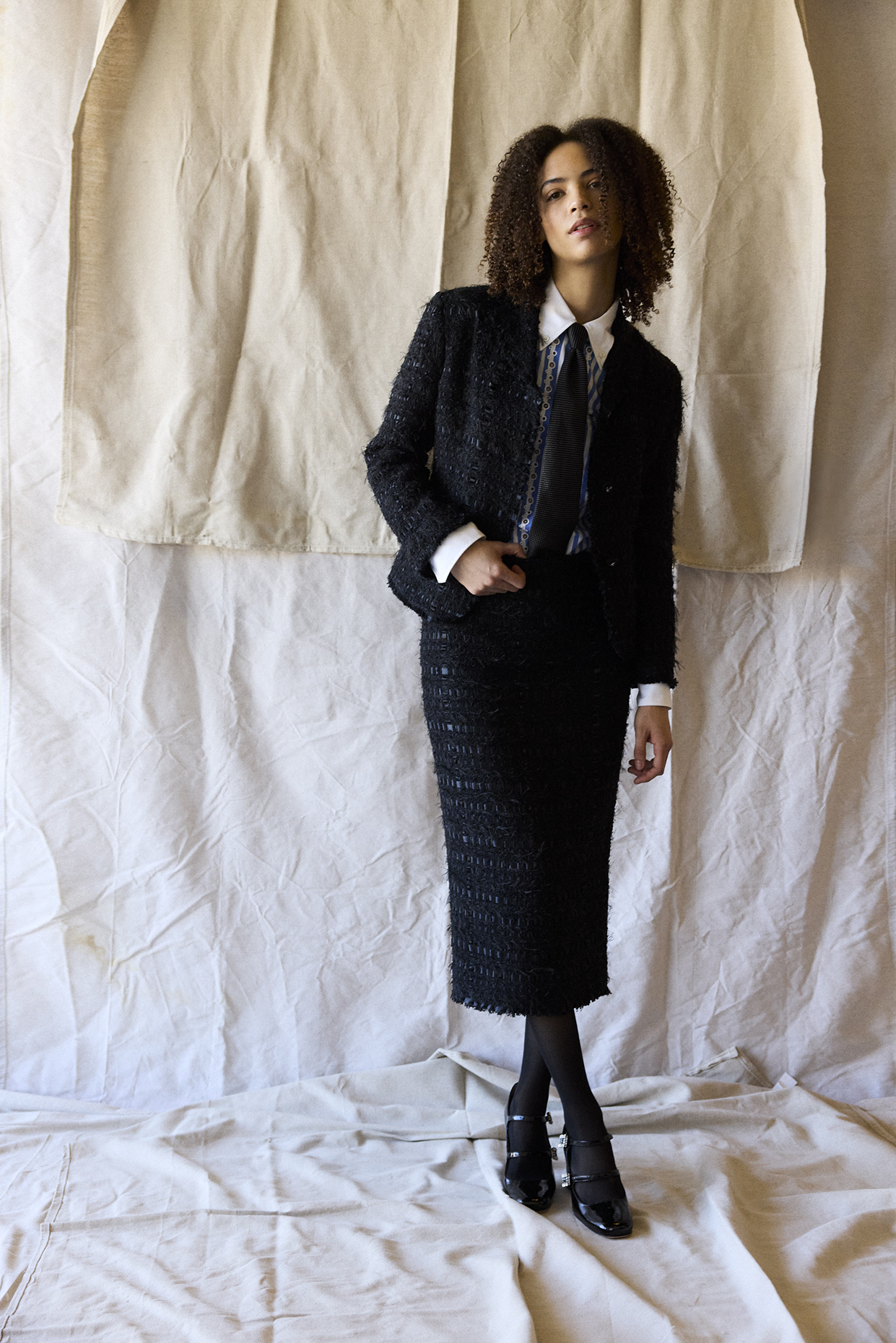 Woman with curly hair in a black textured suit and white shirt, standing against a beige fabric backdrop.