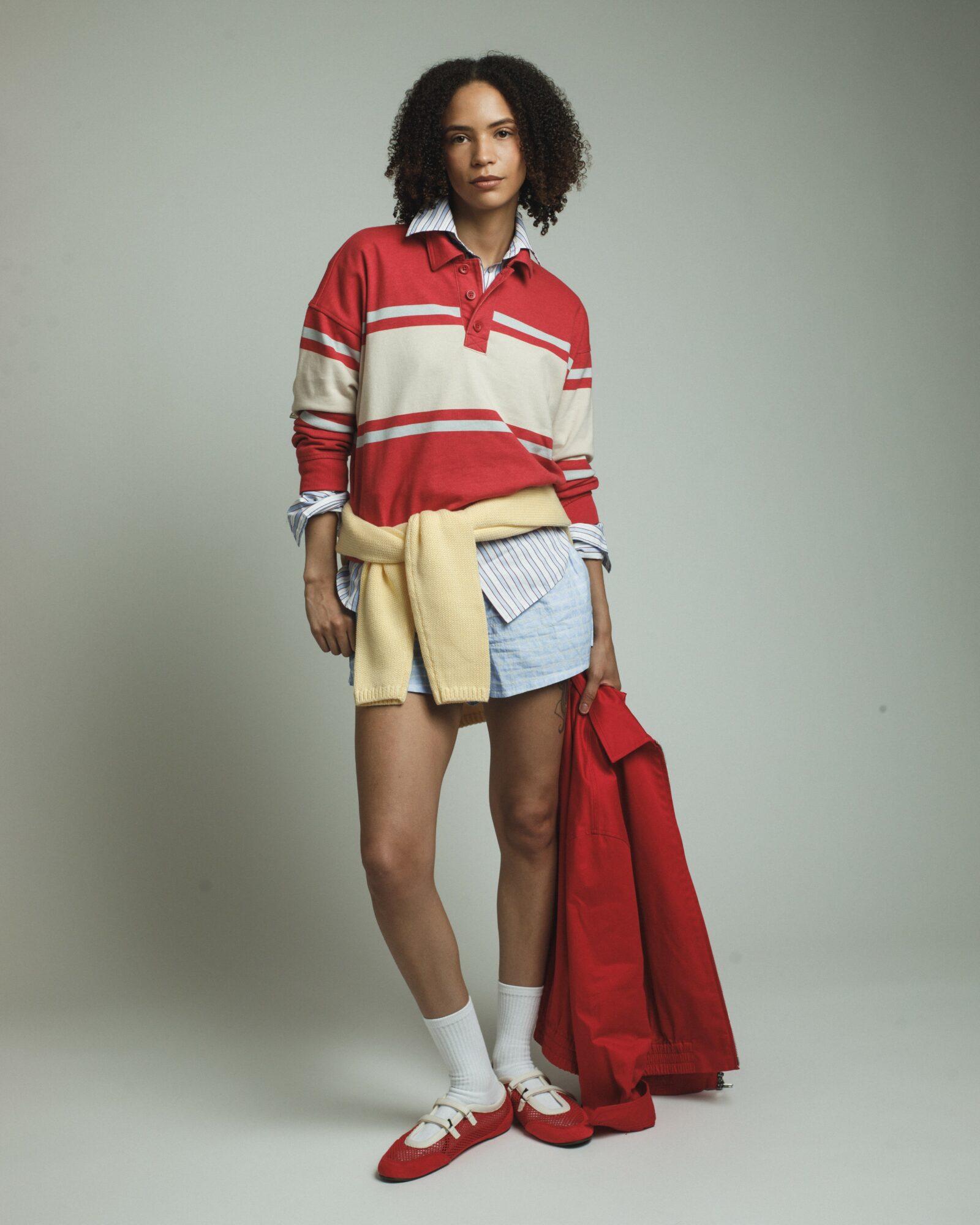 Young woman with curly hair wearing a red and white striped rugby shirt, blue shorts, white socks, and red shoes, holding a red jacket.