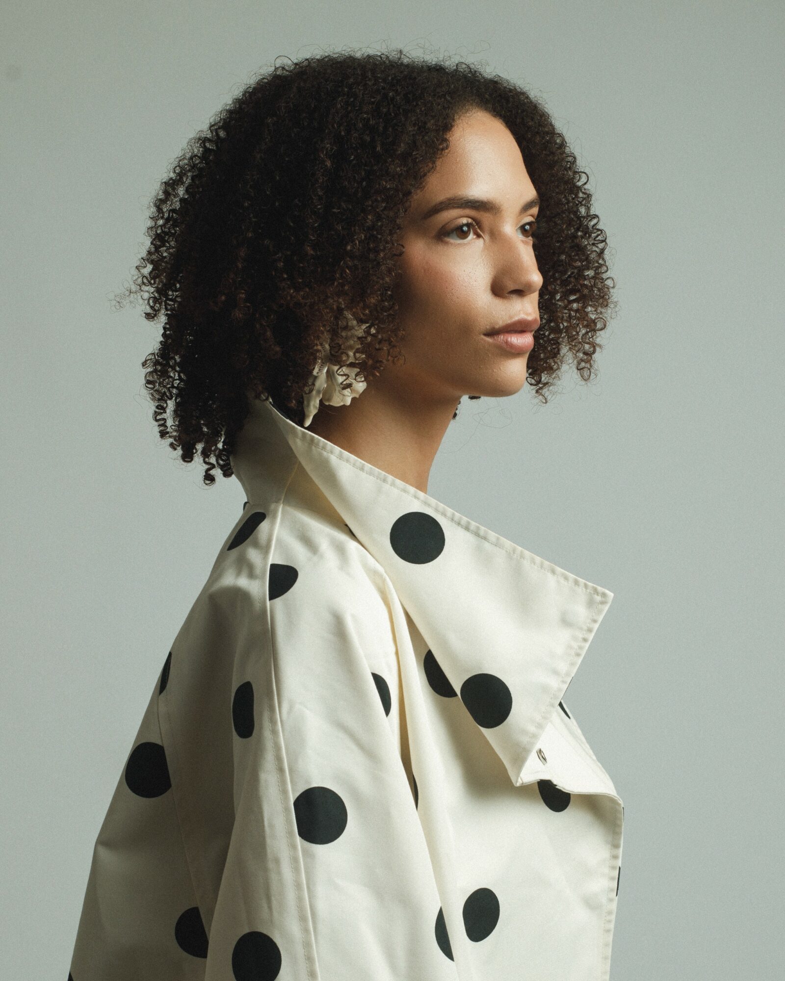 Profile of a woman with curly hair wearing a polka dot jacket against a plain background.