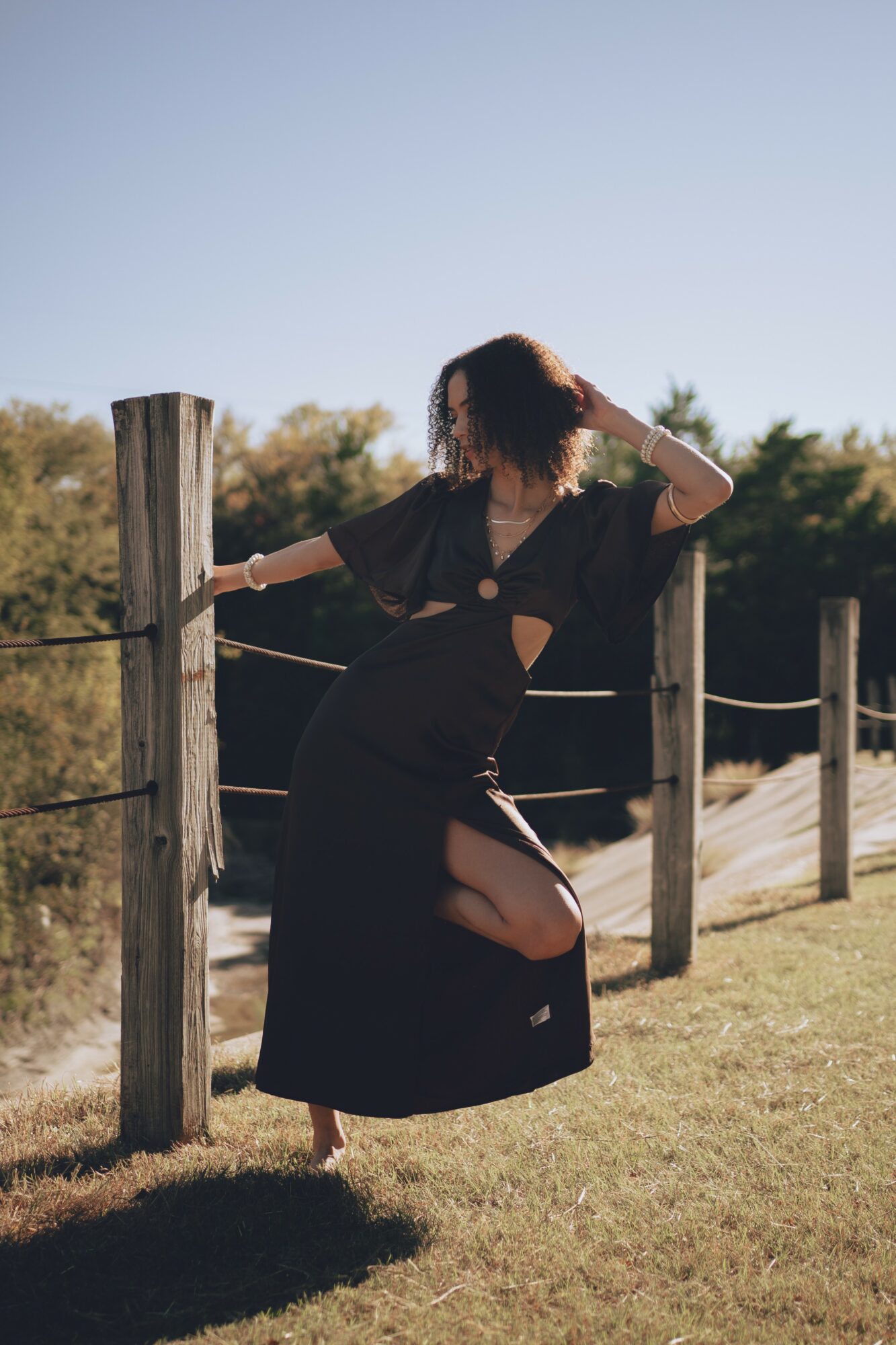 Person standing outdoors near a wooden fence, wearing a black dress with a slit, with trees and sky in background.