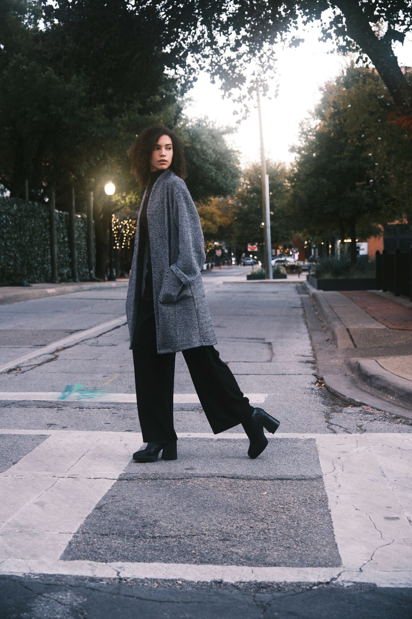 Woman standing on crosswalk in a city street during evening, wearing a gray coat and black pants, trees and streetlights in background.