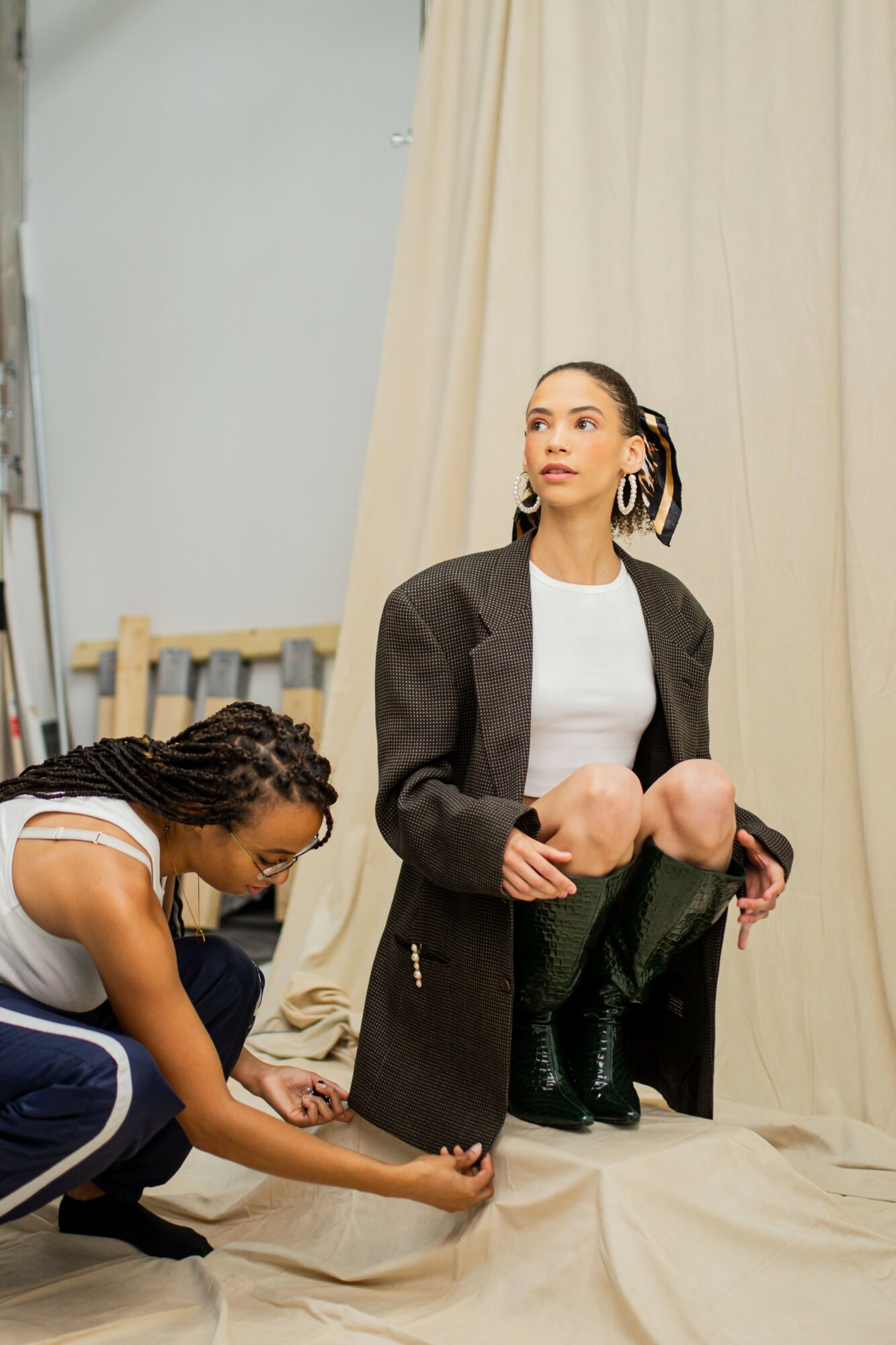 Woman with dark hair in a ponytail, wearing a white shirt and black coat, kneeling with legs crossed, looking upward. Person with braided hair in a white top and blue pants crouching, adjusting a shoe. Beige curtain backdrop.