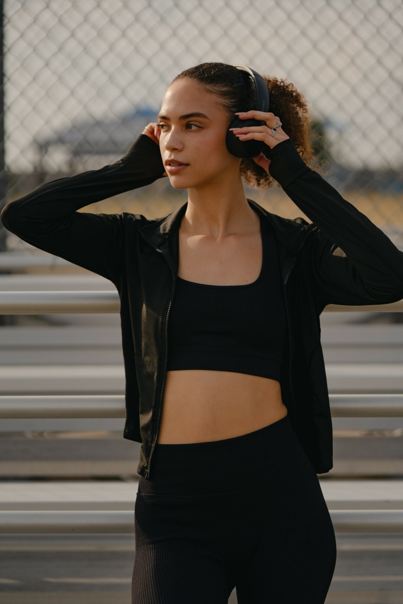 Young woman with dark hair in a ponytail, wearing black athletic clothing, standing outdoors near a fence.