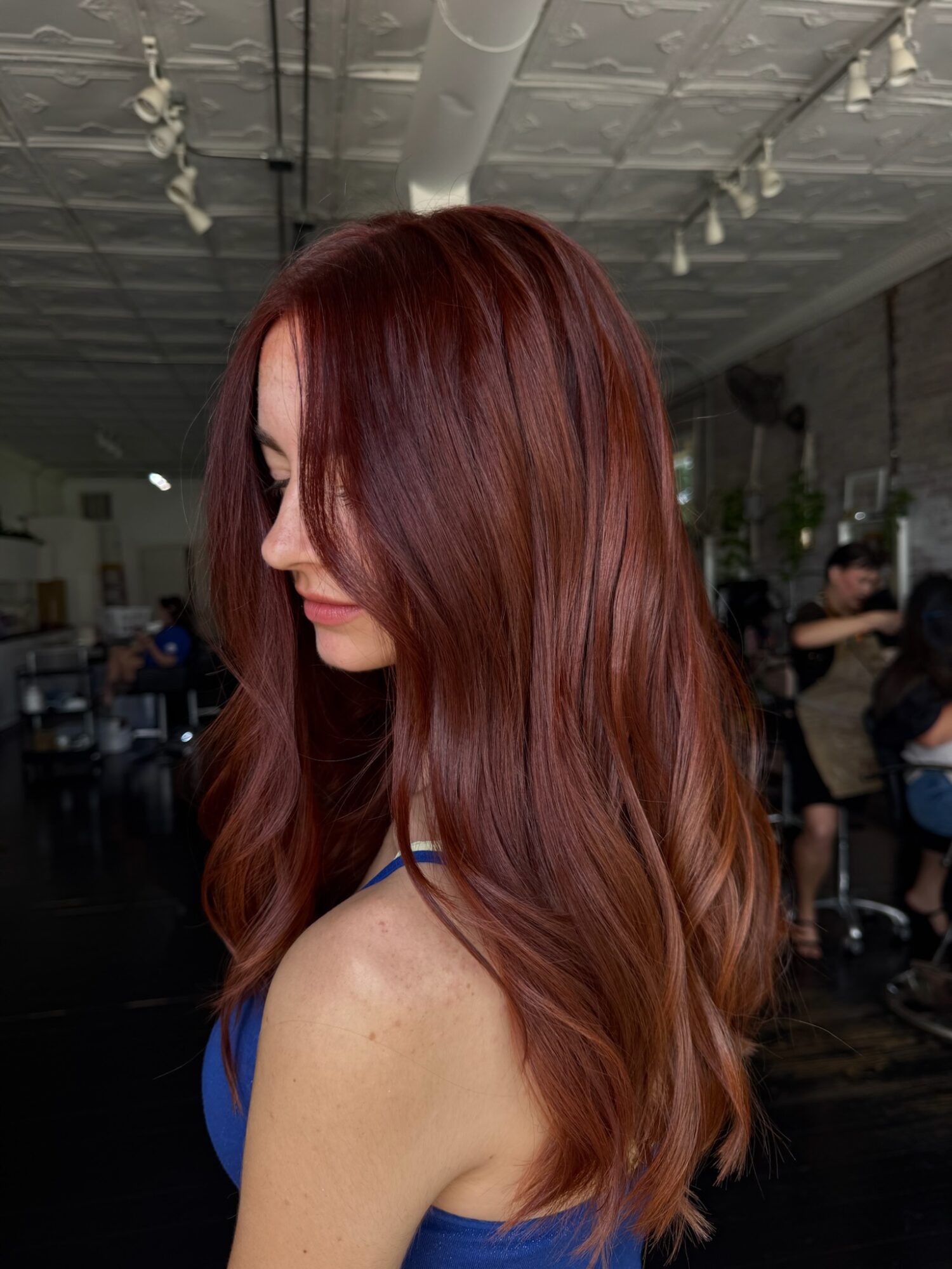 Side view of a woman with long, wavy, reddish-brown hair in an indoor setting.