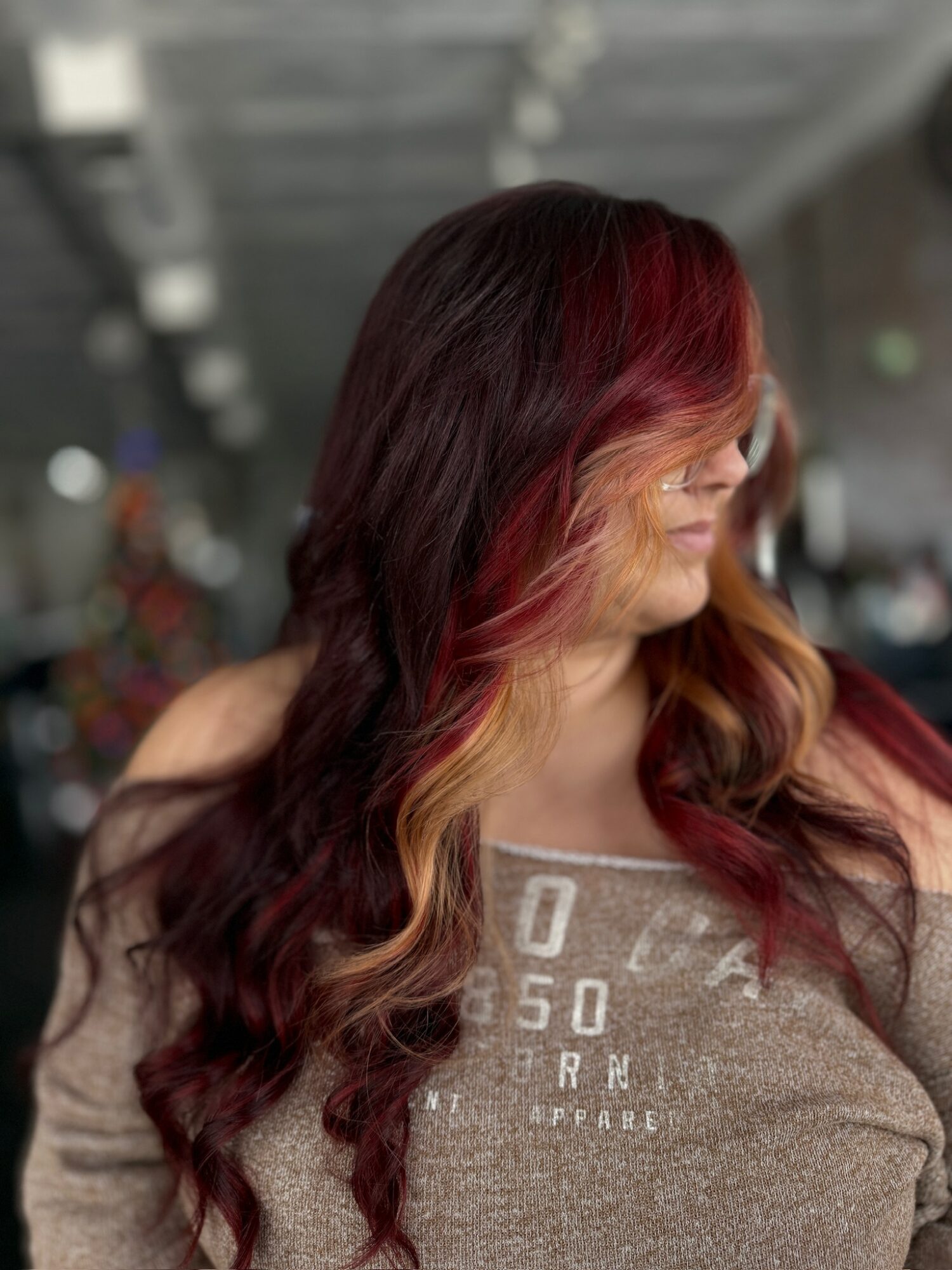 Woman with long, wavy hair dyed in shades of red and burgundy, wearing a gray top, in an indoor setting.