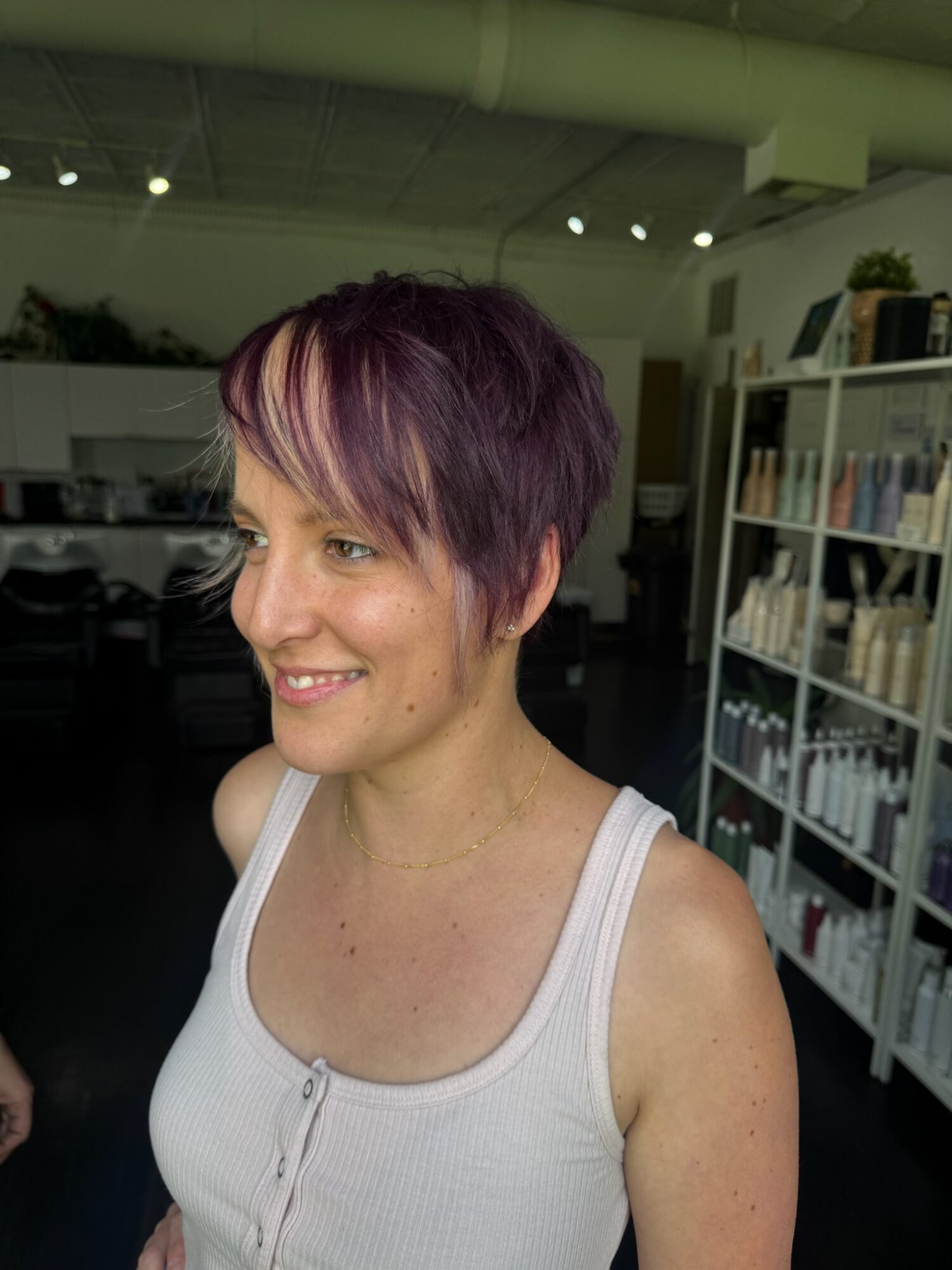 Woman with short purple hair smiling indoors, bookshelf in background.