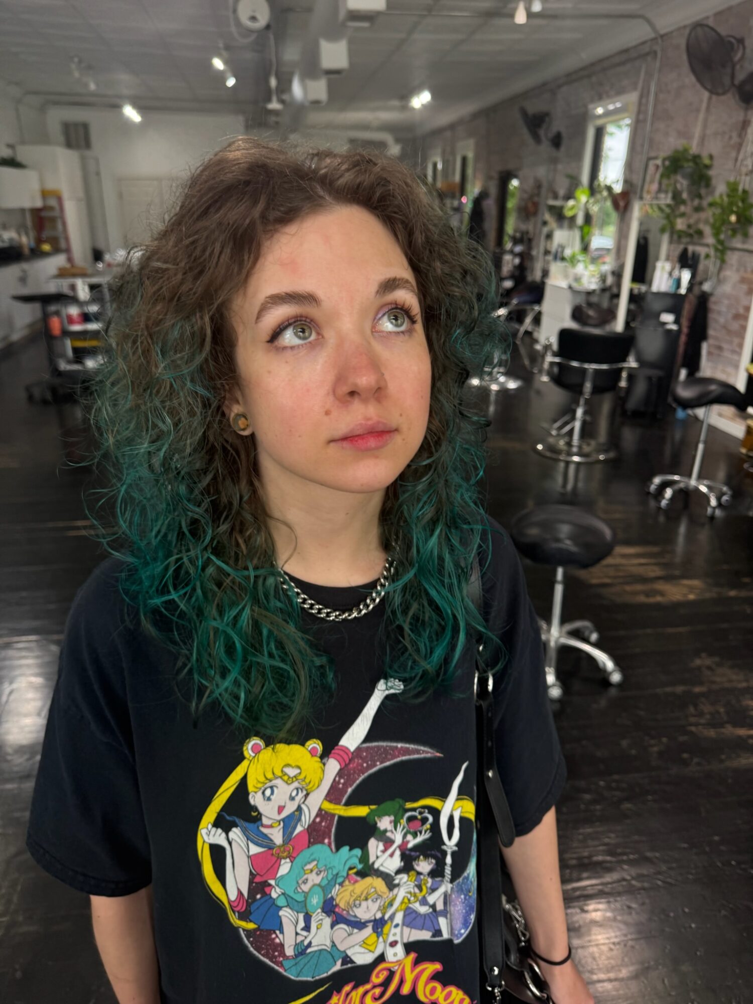 Young woman with curly hair and green tips, wearing a graphic T-shirt, looking upward in a salon, with chairs and plants in background.
