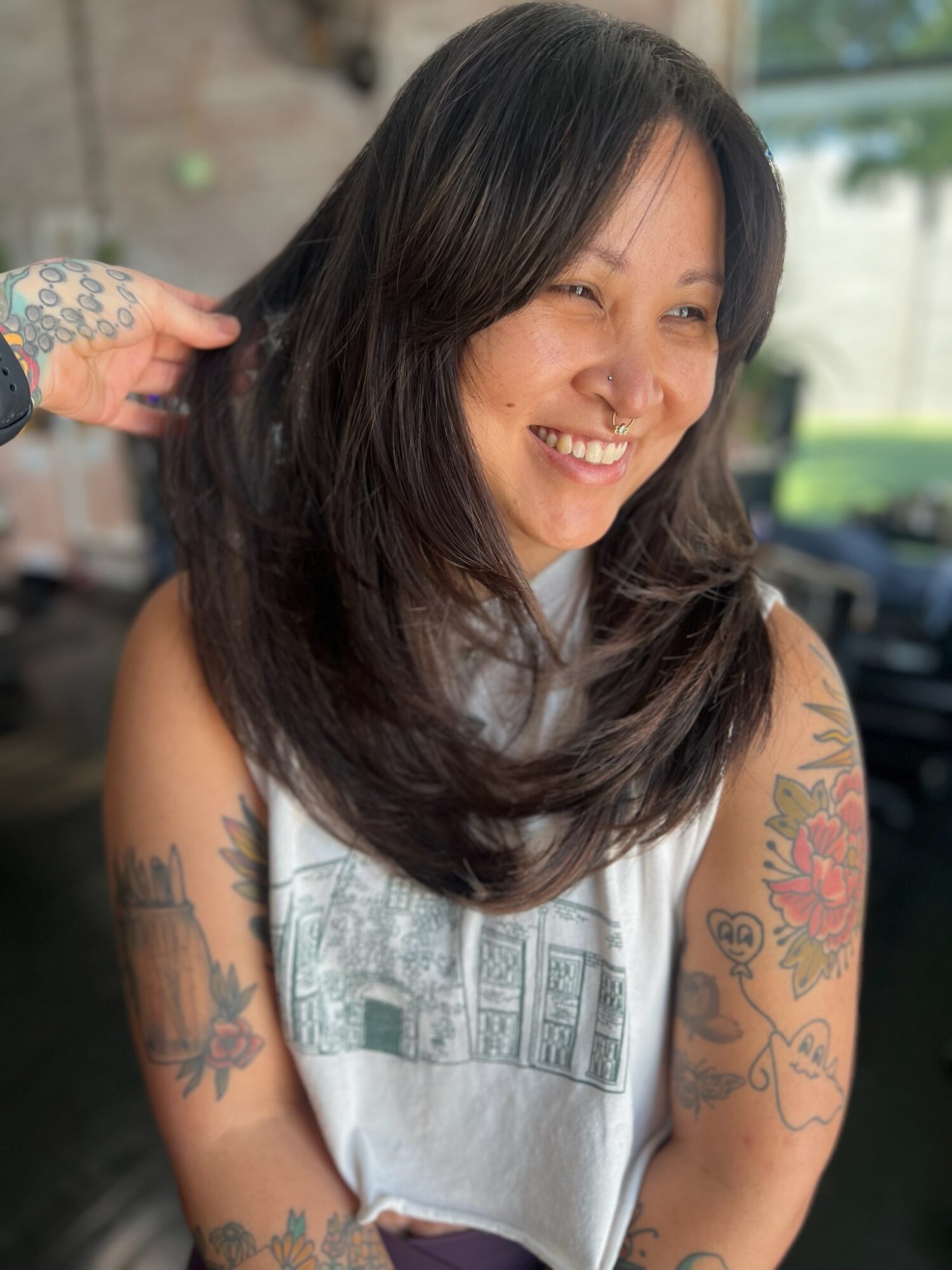 Woman with dark hair smiling, sitting indoors with tattoos on arms, wearing a sleeveless top, background blurred.