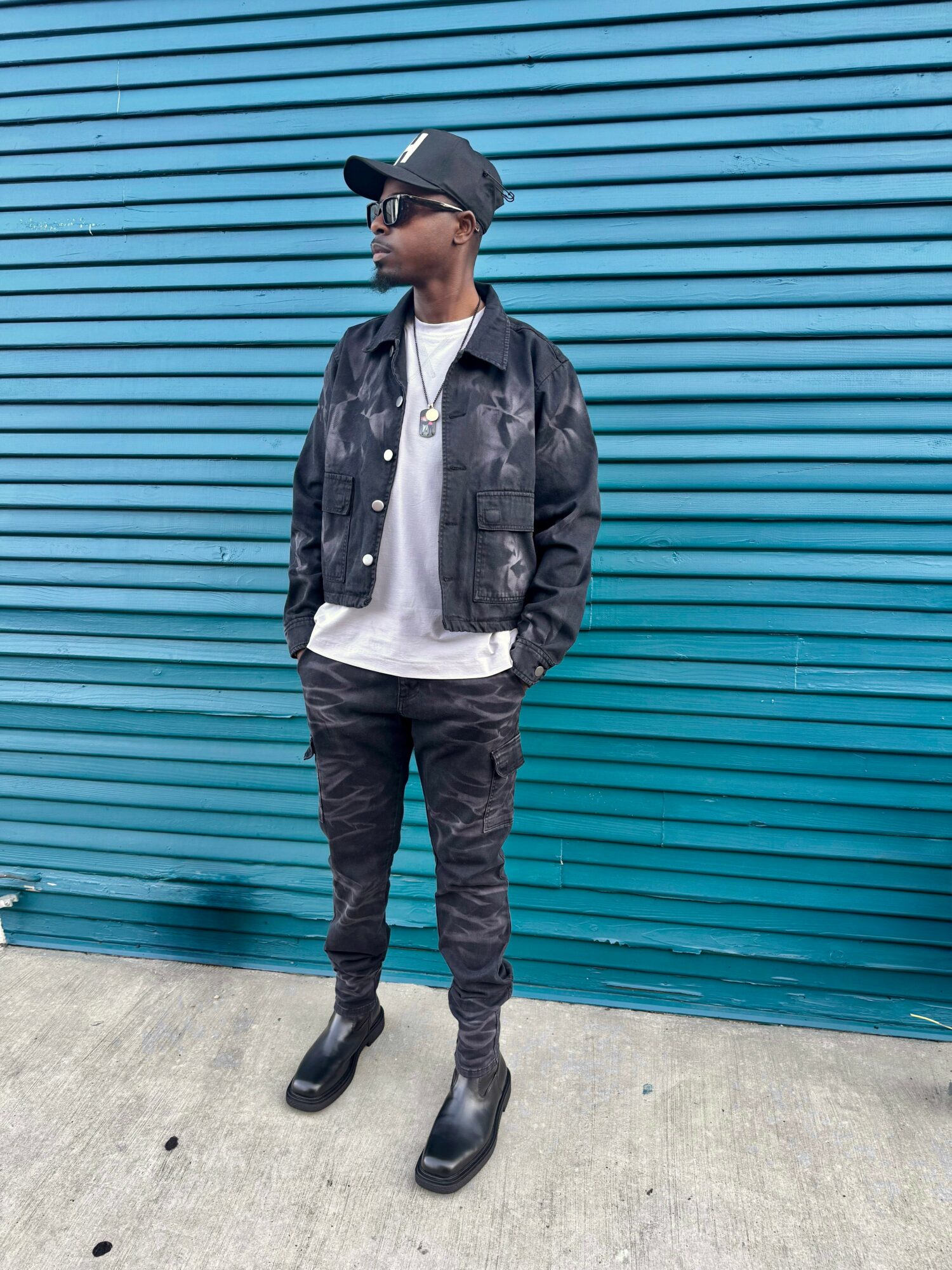 Person standing in front of a blue corrugated metal wall, wearing a black jacket, white shirt, black pants, and boots.
