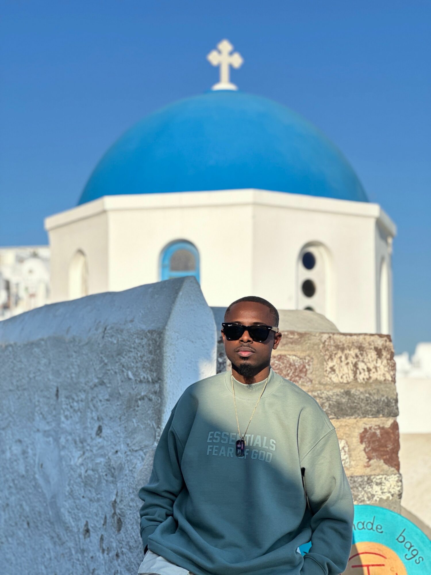 Man wearing sunglasses and a gray sweatshirt standing in front of a white building with a blue dome and cross, clear sky