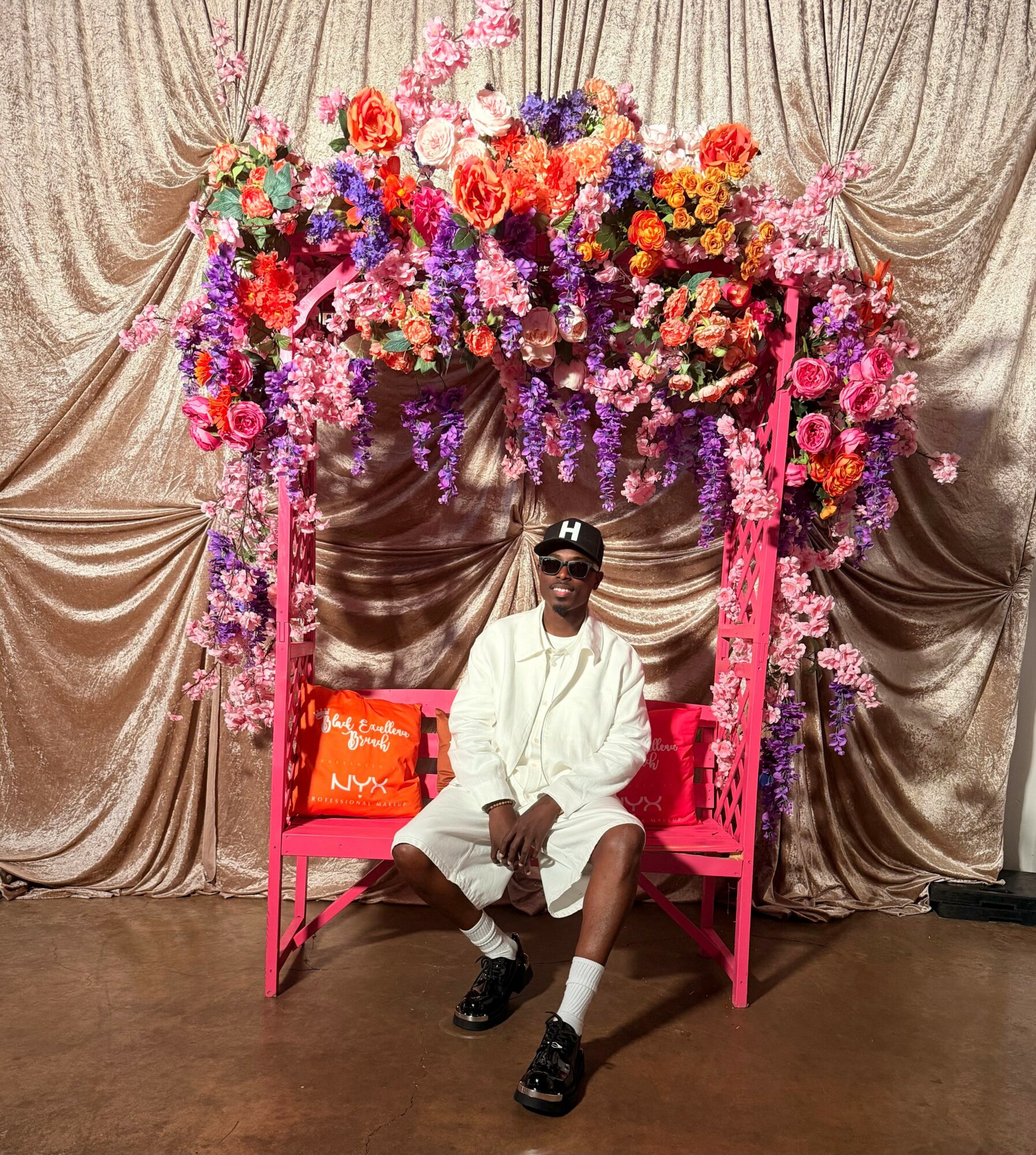 Person sitting on a pink bench under a large floral arrangement with pink, purple, and orange flowers, against a beige curtain backdrop.