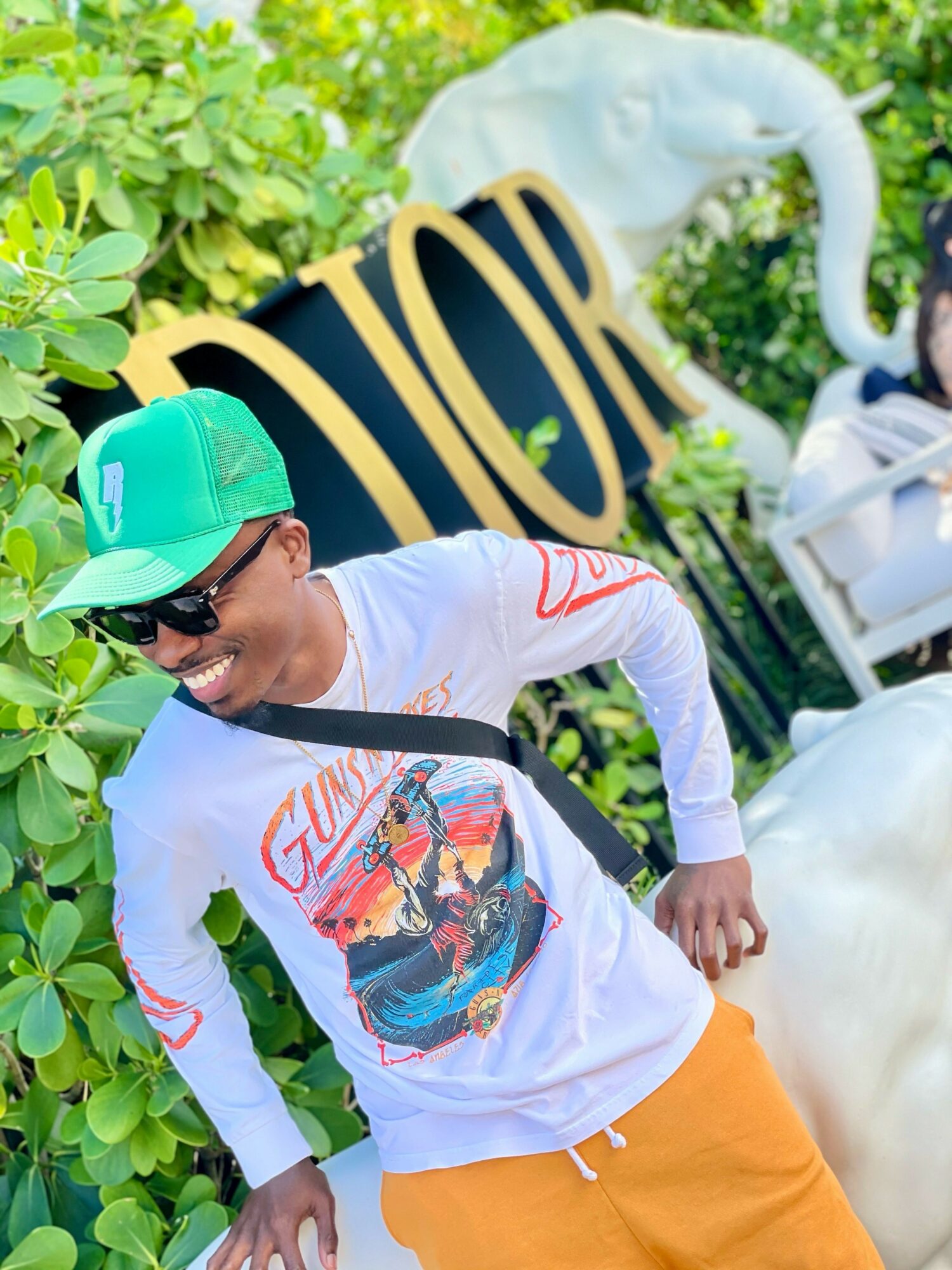 Person smiling, wearing a green cap, sunglasses, white long-sleeve shirt with a colorful graphic, and orange pants, outdoors with greenery and a swan sculpture.