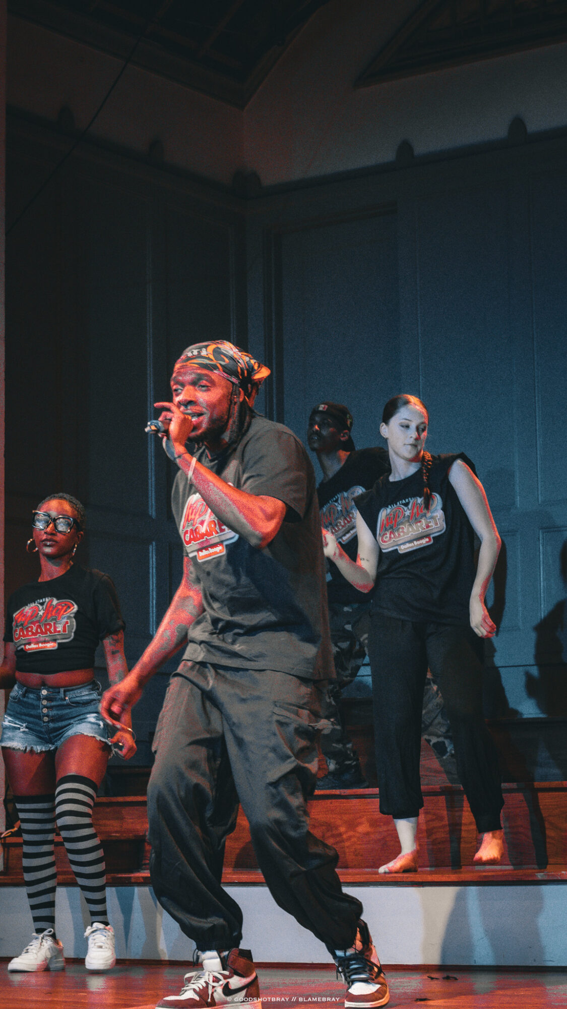 Four performers on stage, one singing into a microphone, others standing and watching, in a dark room with blue walls.